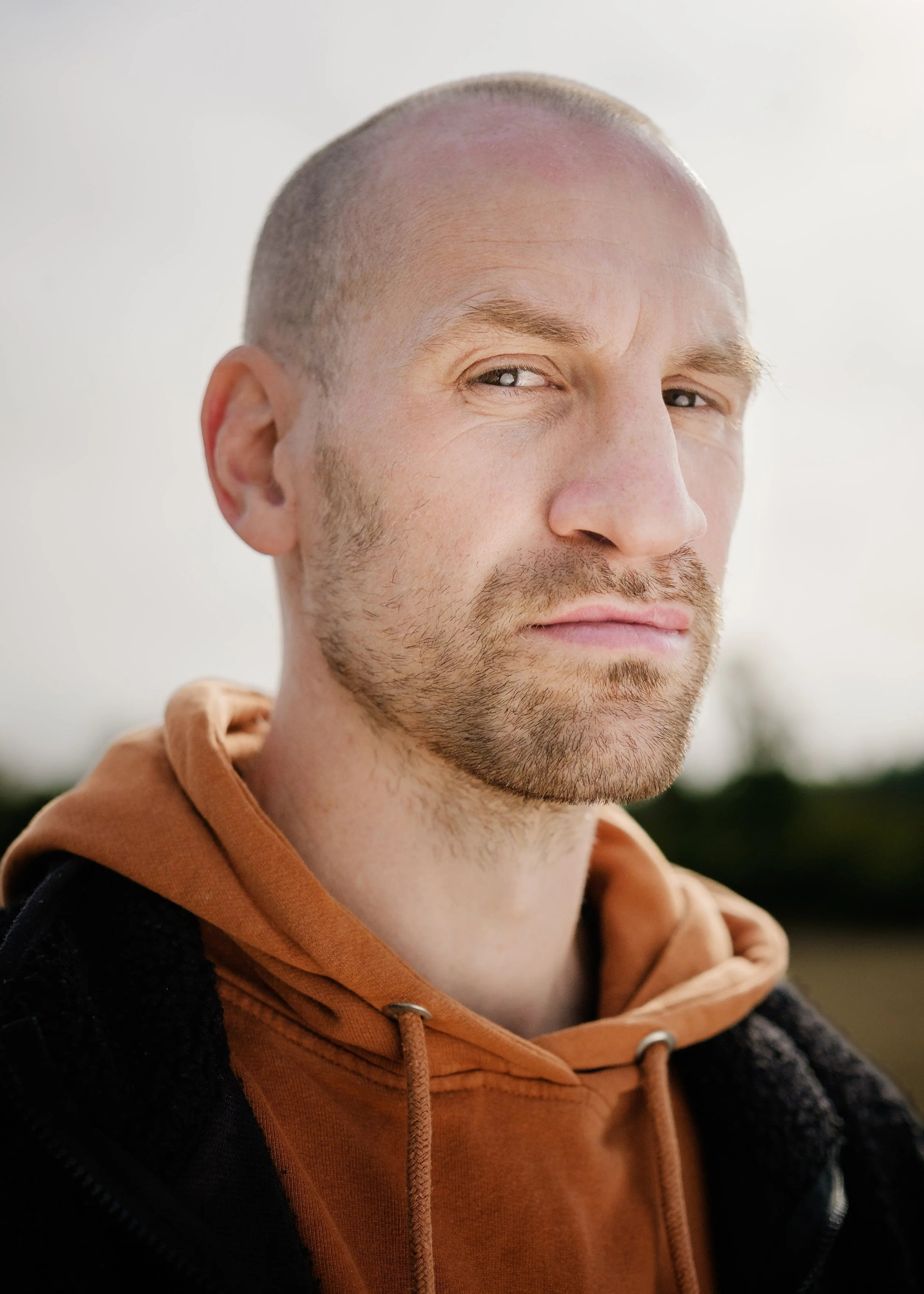 Porträtt av en man med skägg, blondtitel, och lätt rakat huvud, rundad haka, iförd en orange hoodie och svart jacka, utomhus.