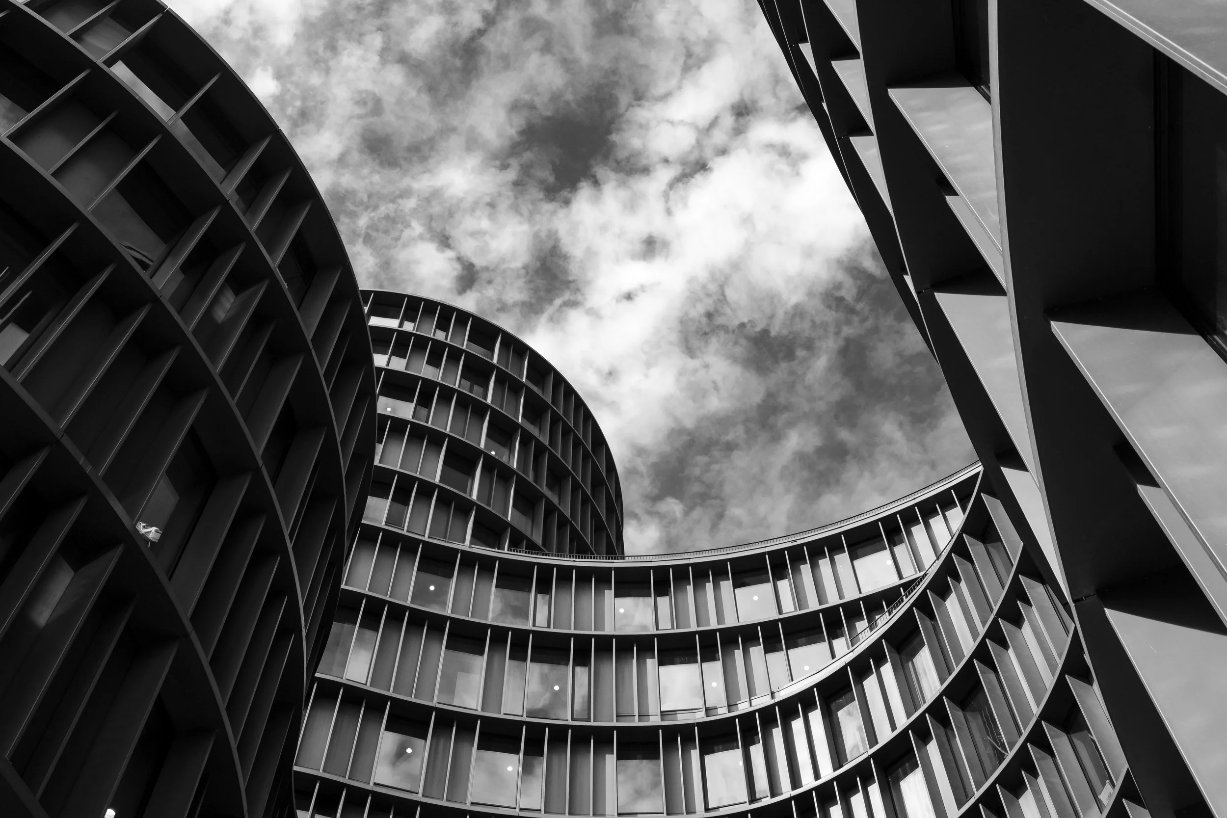 Svartvit bild av moderna höga byggnader med rundade och kubiska former sett från marknivå mot en skyfull himmel.