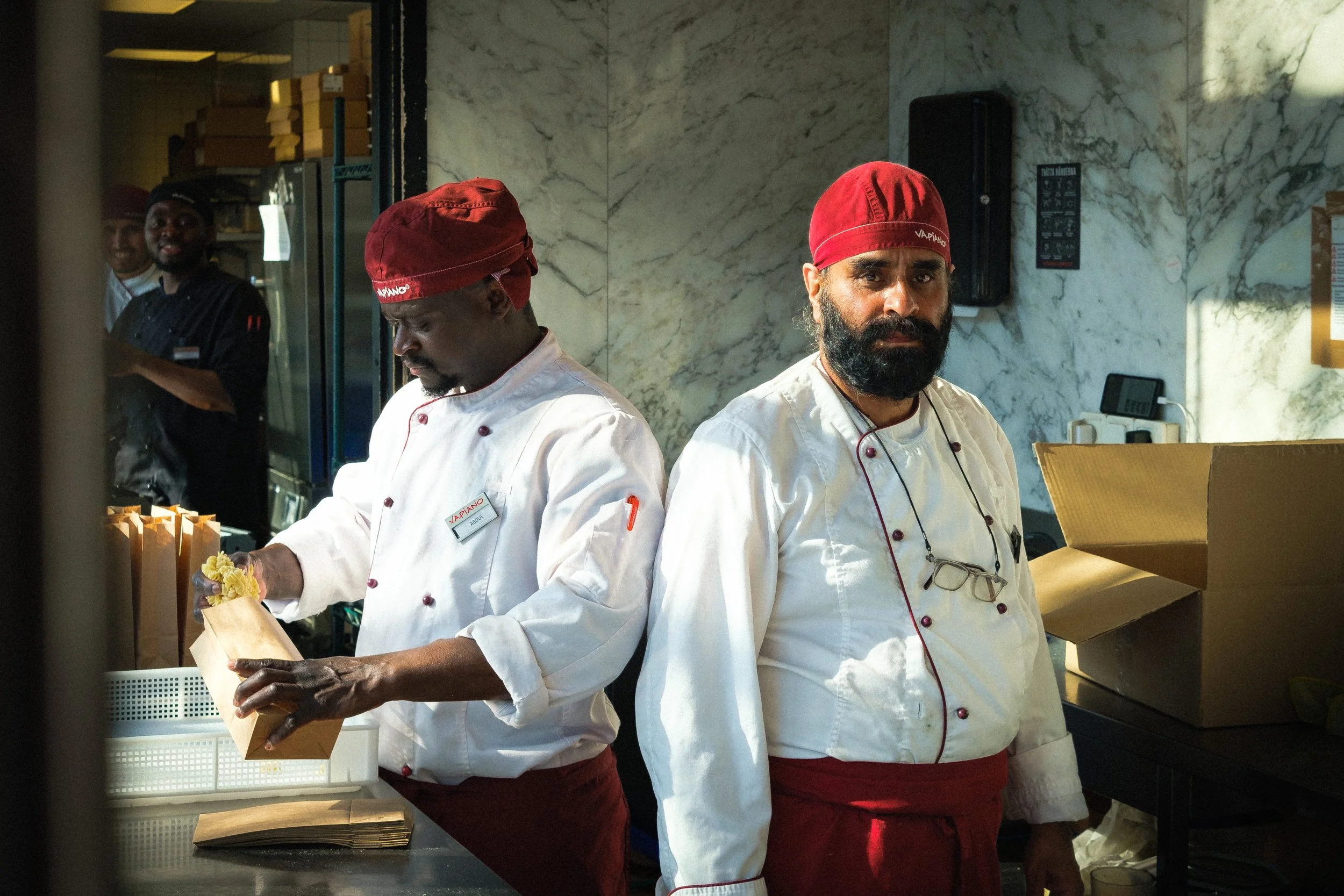 Två kockar i köket, en som packar ett papper och en som tittar mot kameran, båda i vita kockkläder och röda kockmössor, i ett kök med marmorskivor på väggen.