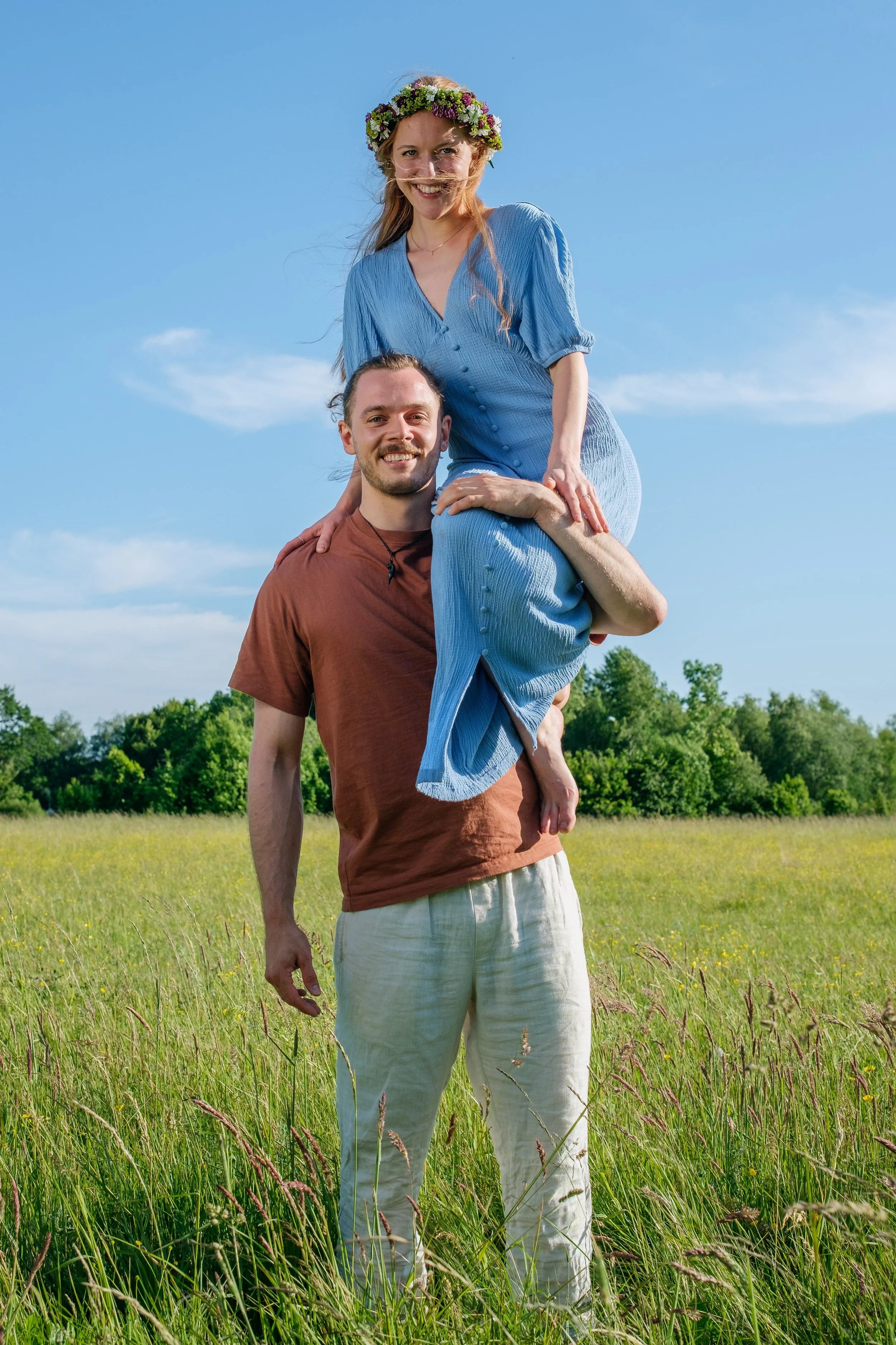 En man som bär en kvinna på axeln i ett grönt fält med träd i bakgrunden och en blå himmel.