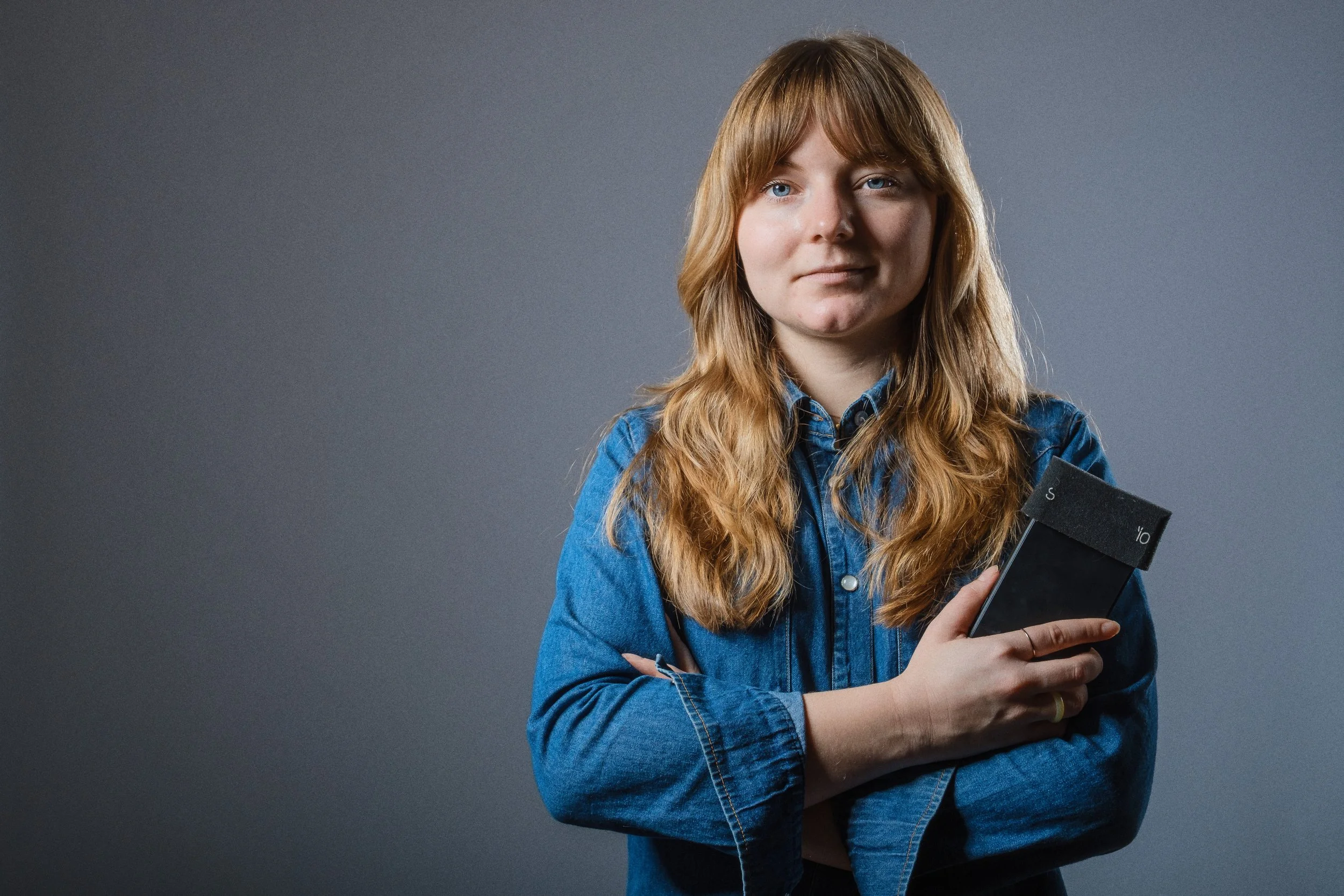 Ung kvinna med rödbrunt, lockigt hår, iklädd jeansjacka, håller i en smartphone mot en grå bakgrund.