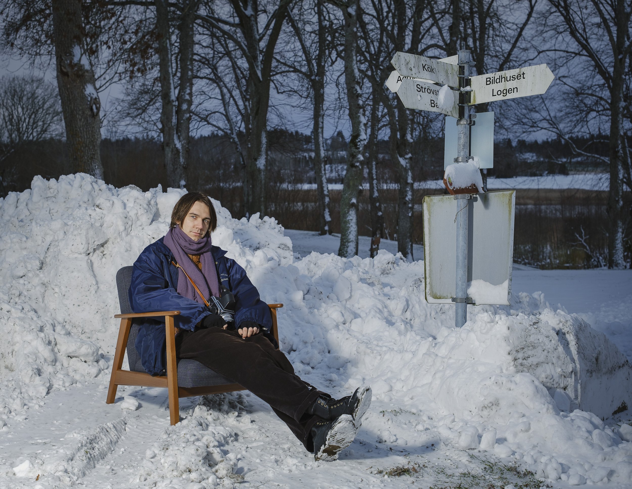 En person sitter i en stol i snön, omgiven av snöiga träd och en vägskylt med flera riktningar märkt med svenska ord, på en vinterdag.