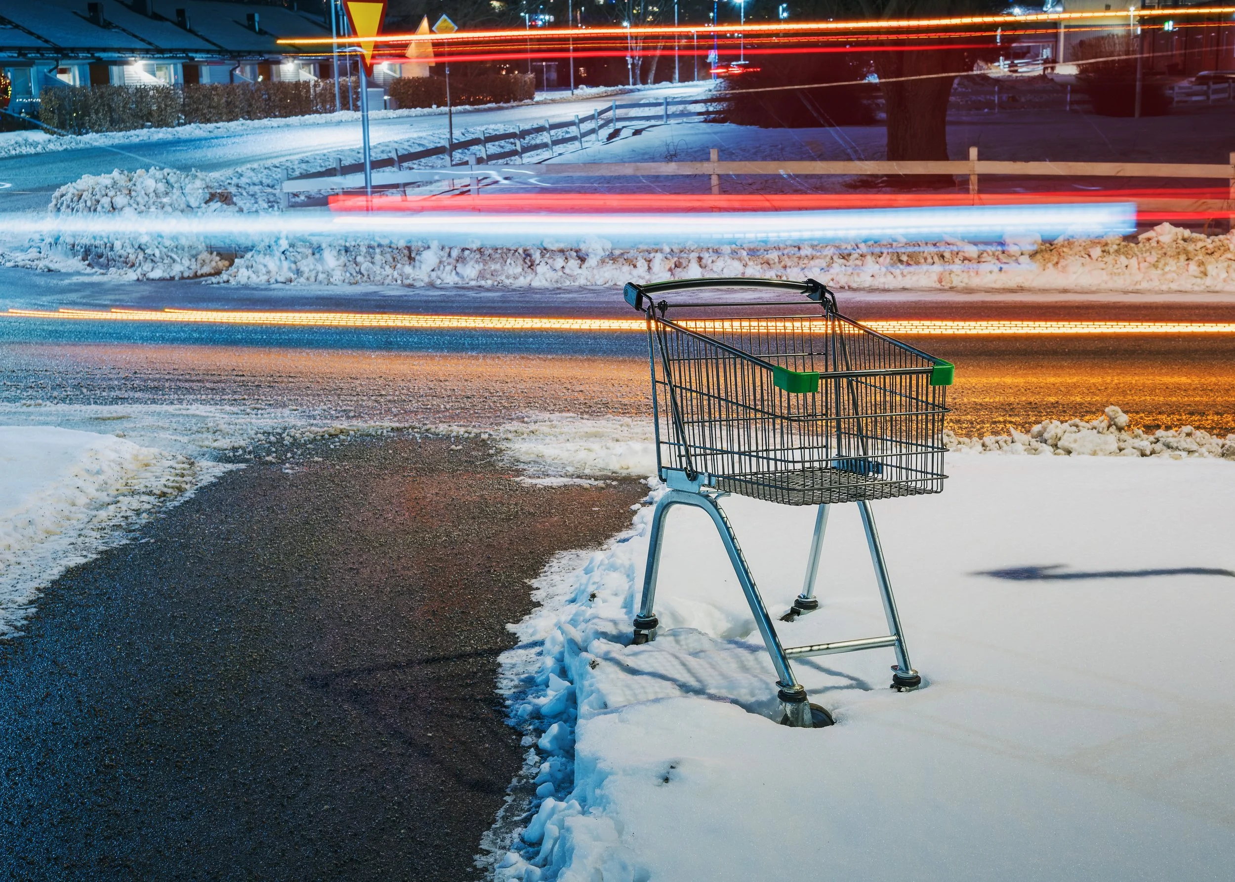 En tom shoppingvagn står på snötäckta marken utanför en affär, med trafik och ljusspår i bakgrunden på kvällen.
