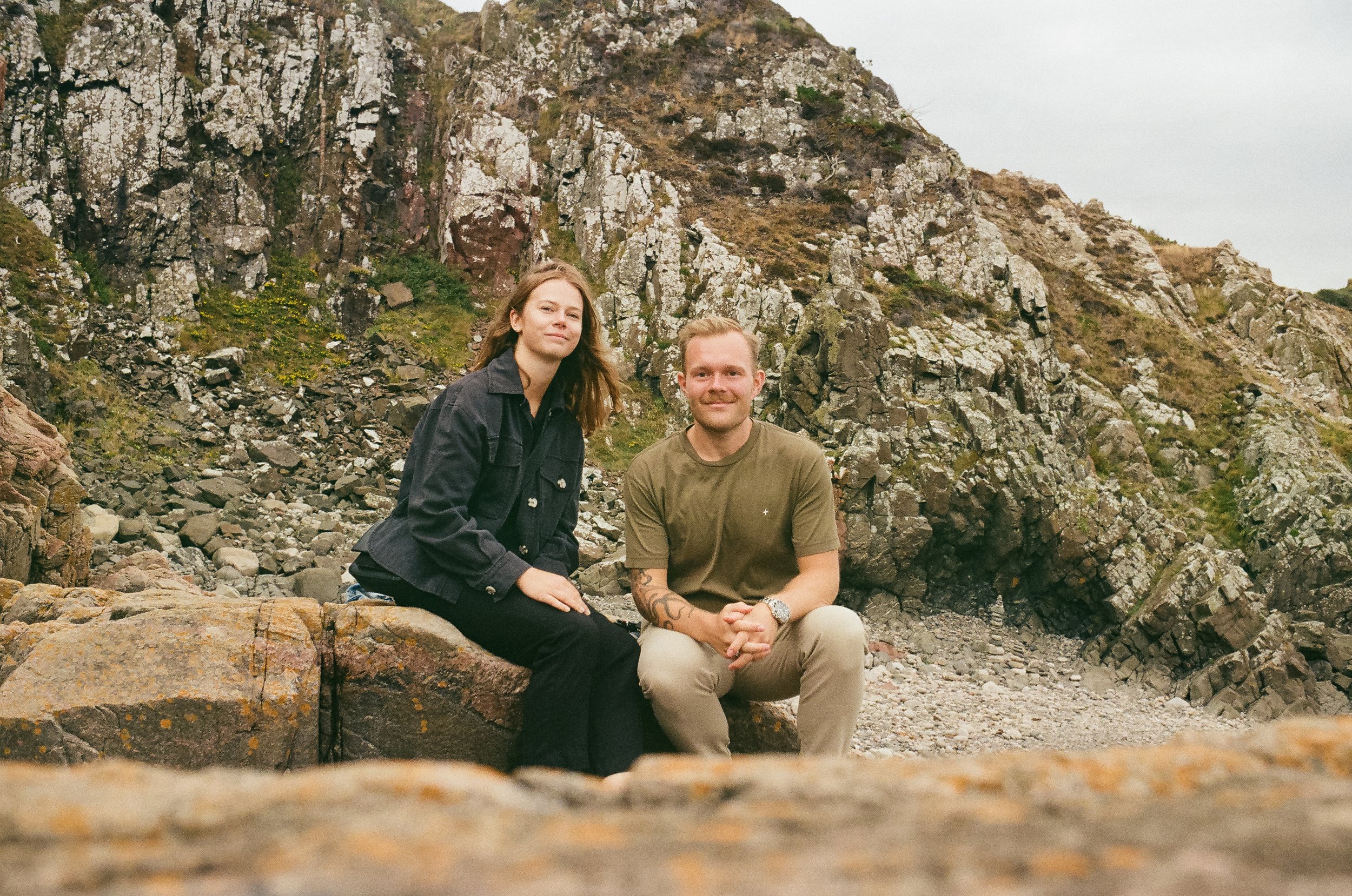 Två personer, en man och en kvinna, sitter på en sten framför en bergig landskap med klippor och gräs i bakgrunden.