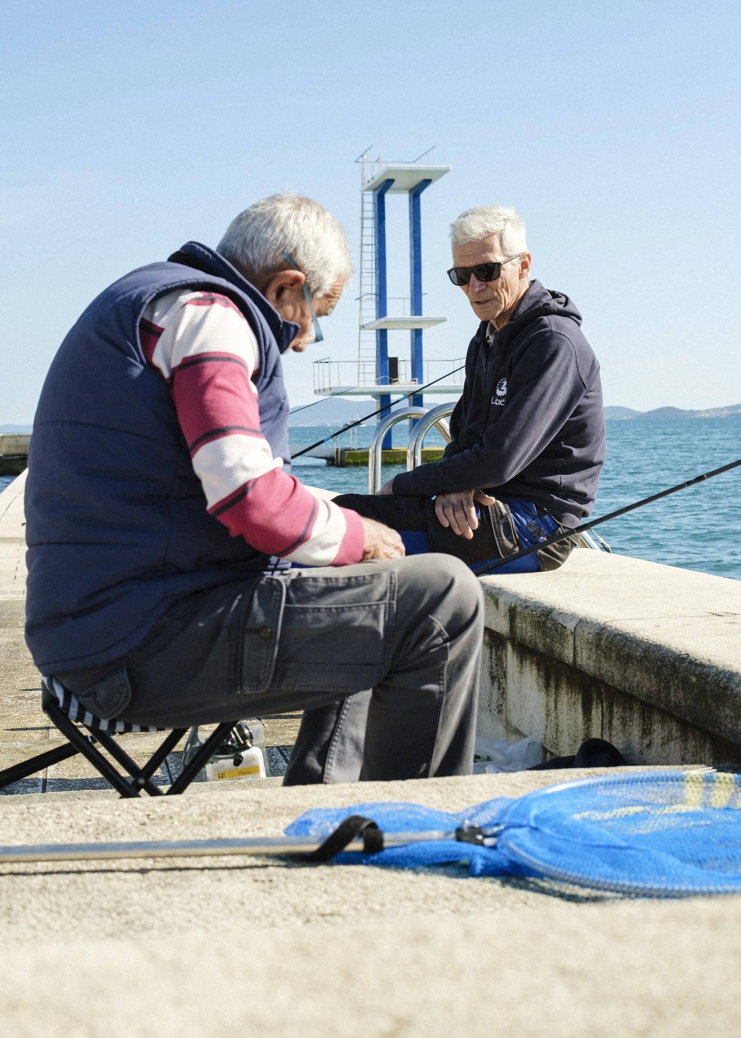 Två äldre män fiskar vid en kaj vid havet, en människa har på sig solglasögon och en annan skriver eller rensar fisk.