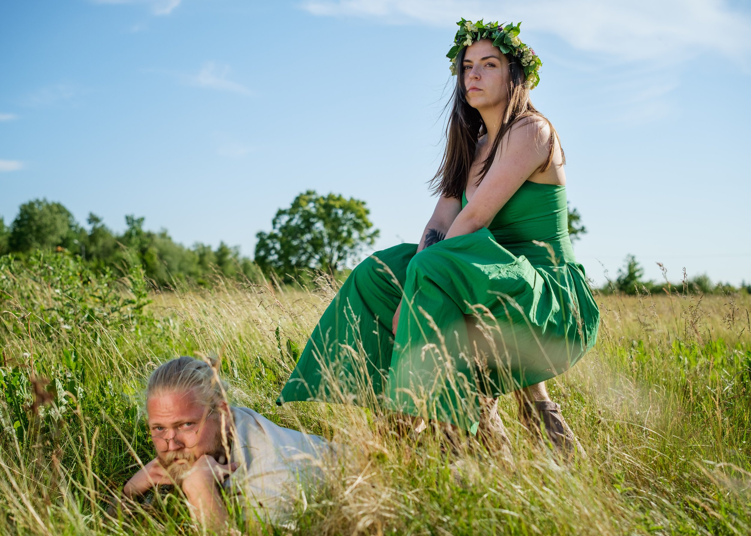 En kvinna i grönt samtidigt som hon bär en blomsterkrans, sitter i ett fält med gräs, medan en man med glasögon ligger på marken under henne.