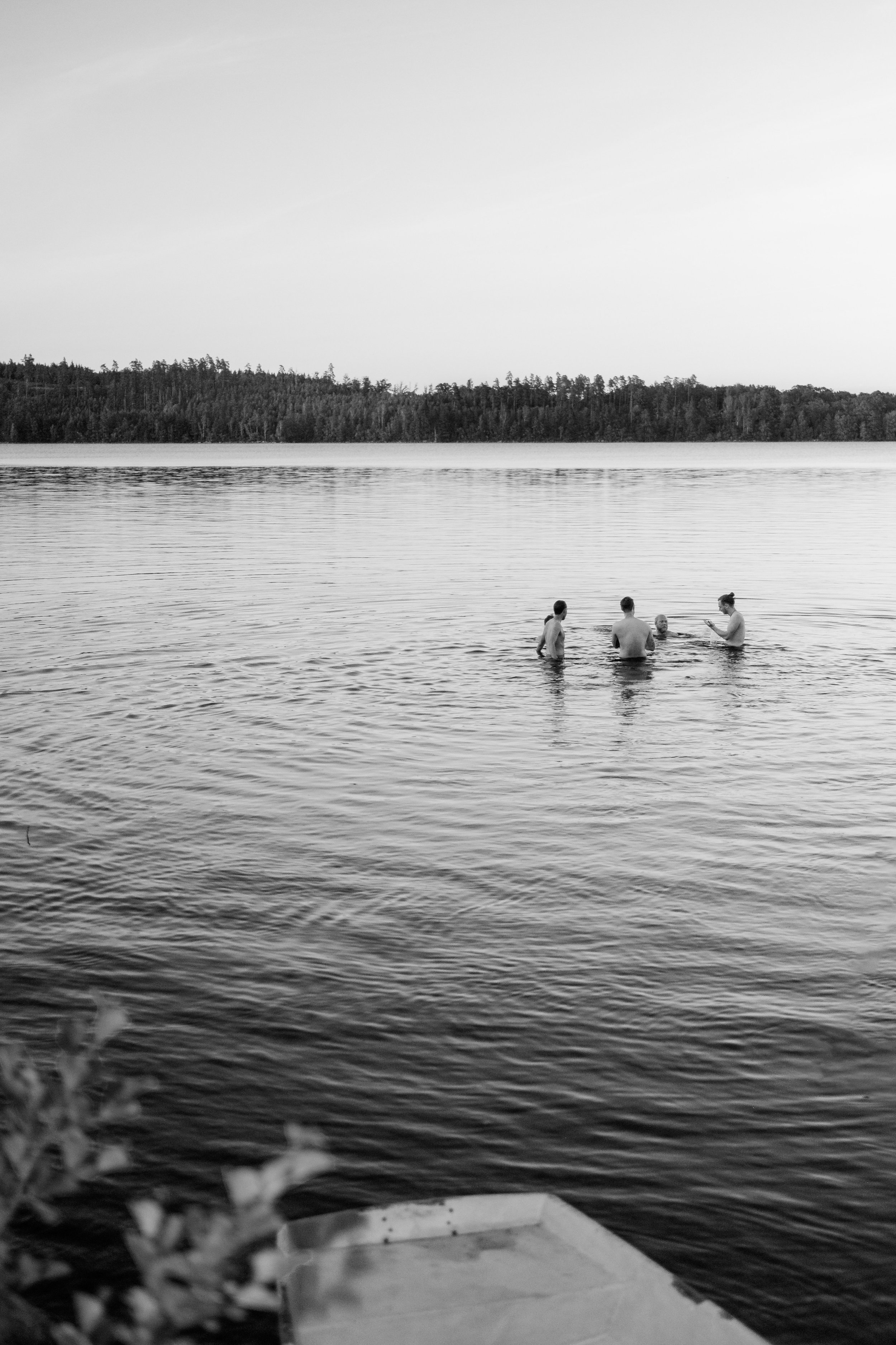 Fyra personer i vattnet vid en sjö, med träd och himmel i bakgrunden, i svartvitt.