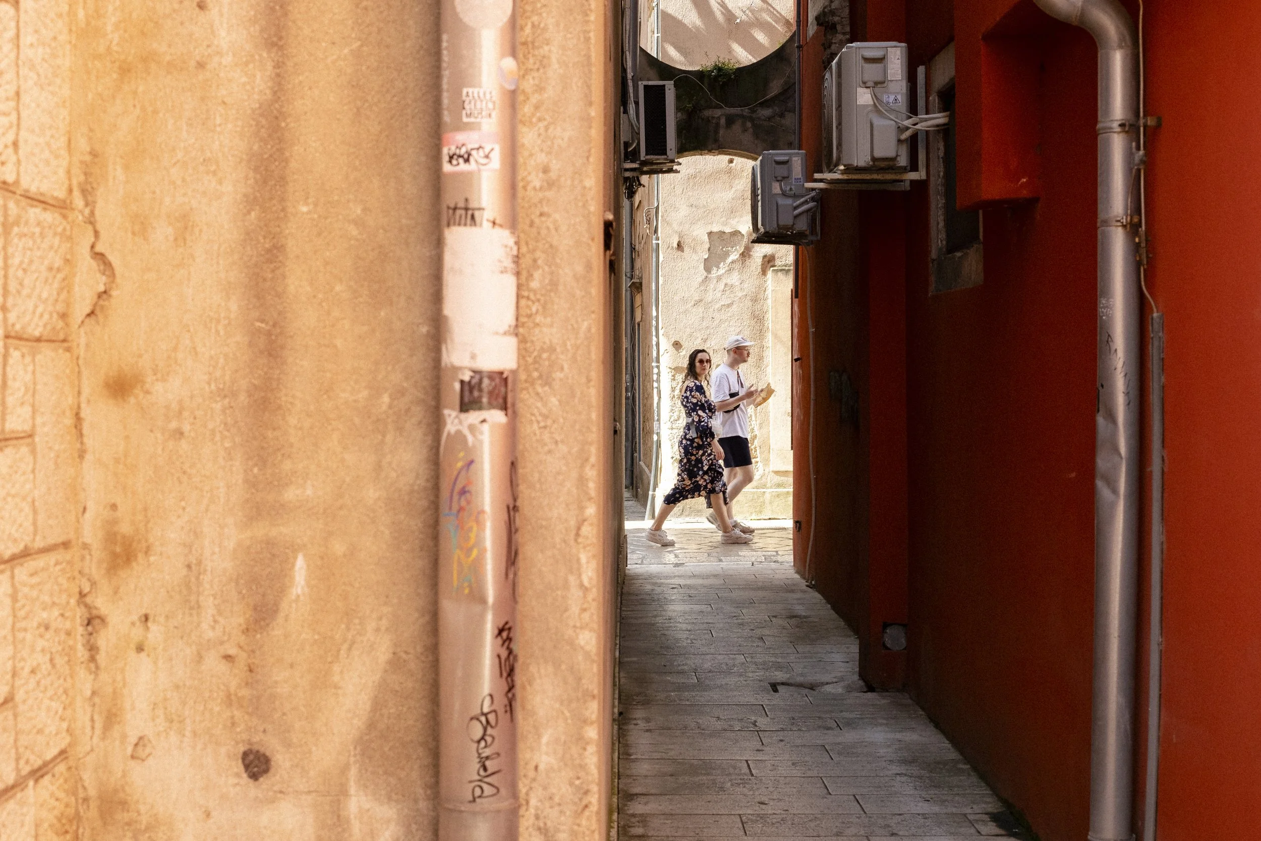 Två personer går igenom en smal gränd, en person håller i en karta och båda bär solglasögon. De är omgivna av väggar i jordfärger, en med graffiti.