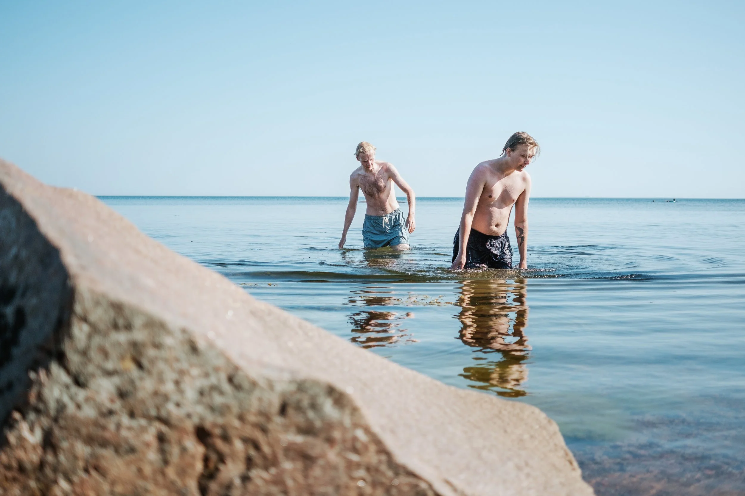 Två unga män som går ut ur havet, klädda i badshorts, med en sandig strand och klar blå himmel i bakgrunden.