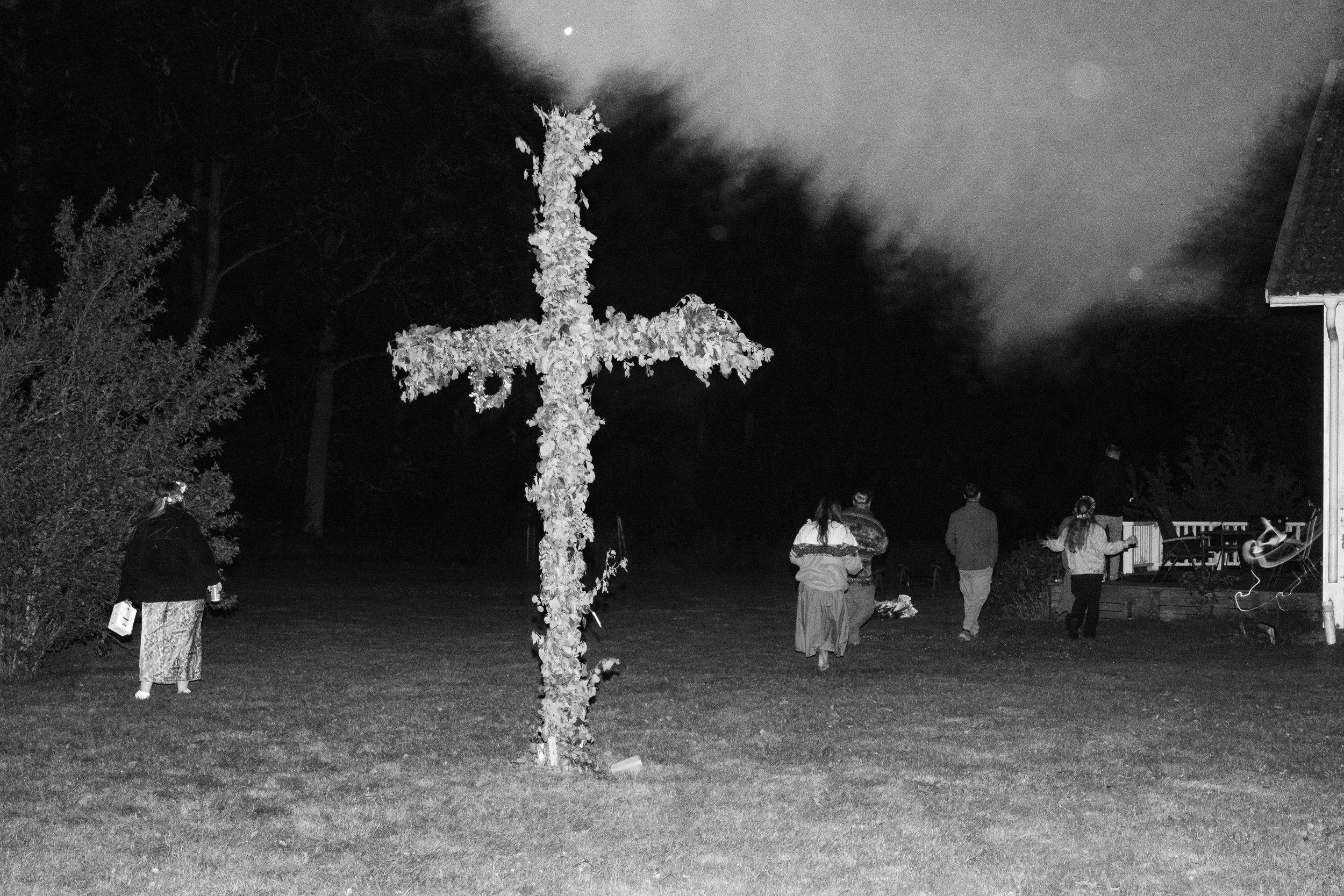 En grupp personer går mot en eldstad i form av ett kors som är täckt av blommor, på natten.