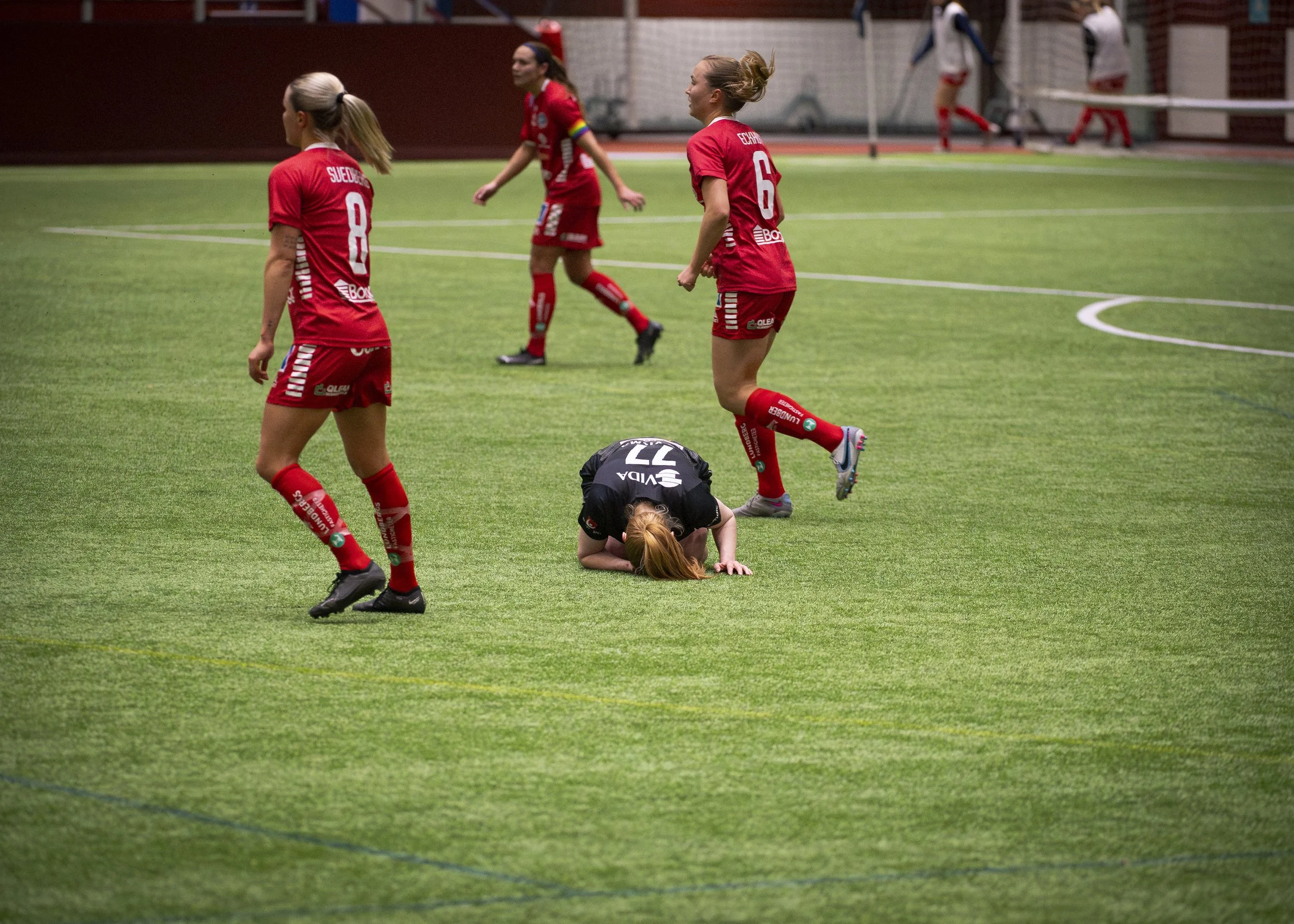 En kvinna i svart träningskläder ligger på knä på planen med huvudet neråt medan andra kvinnor i röda fotbollsspelarkläder står och går runt henne. Det ser ut som en skada eller ett olycksögonblick under en fotbollsmatch.