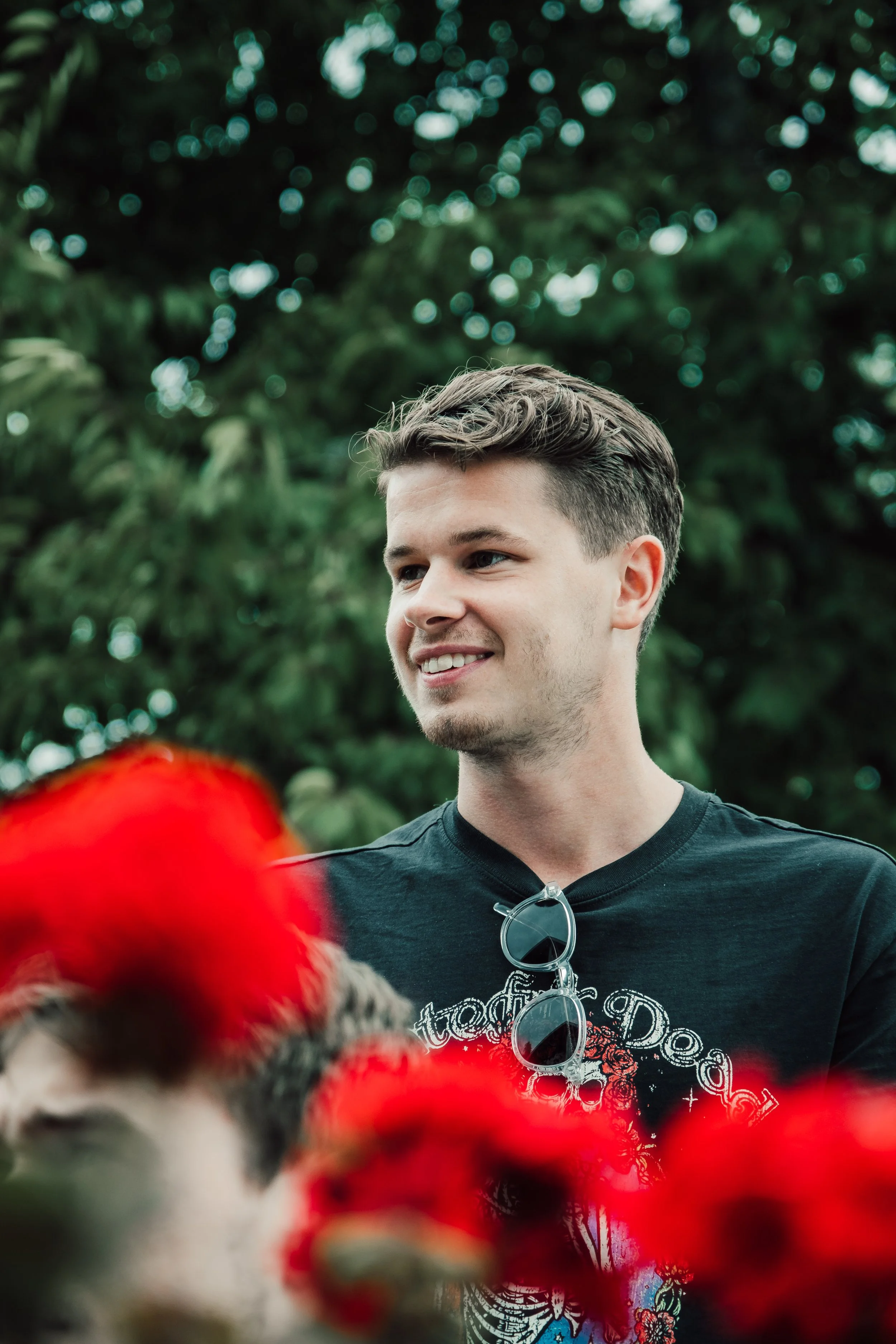 En ung man med kort, brunt hår, som ler och tittar åt sidan, utomhus, med gröna träd i bakgrunden och röda blommor i förgrunden.