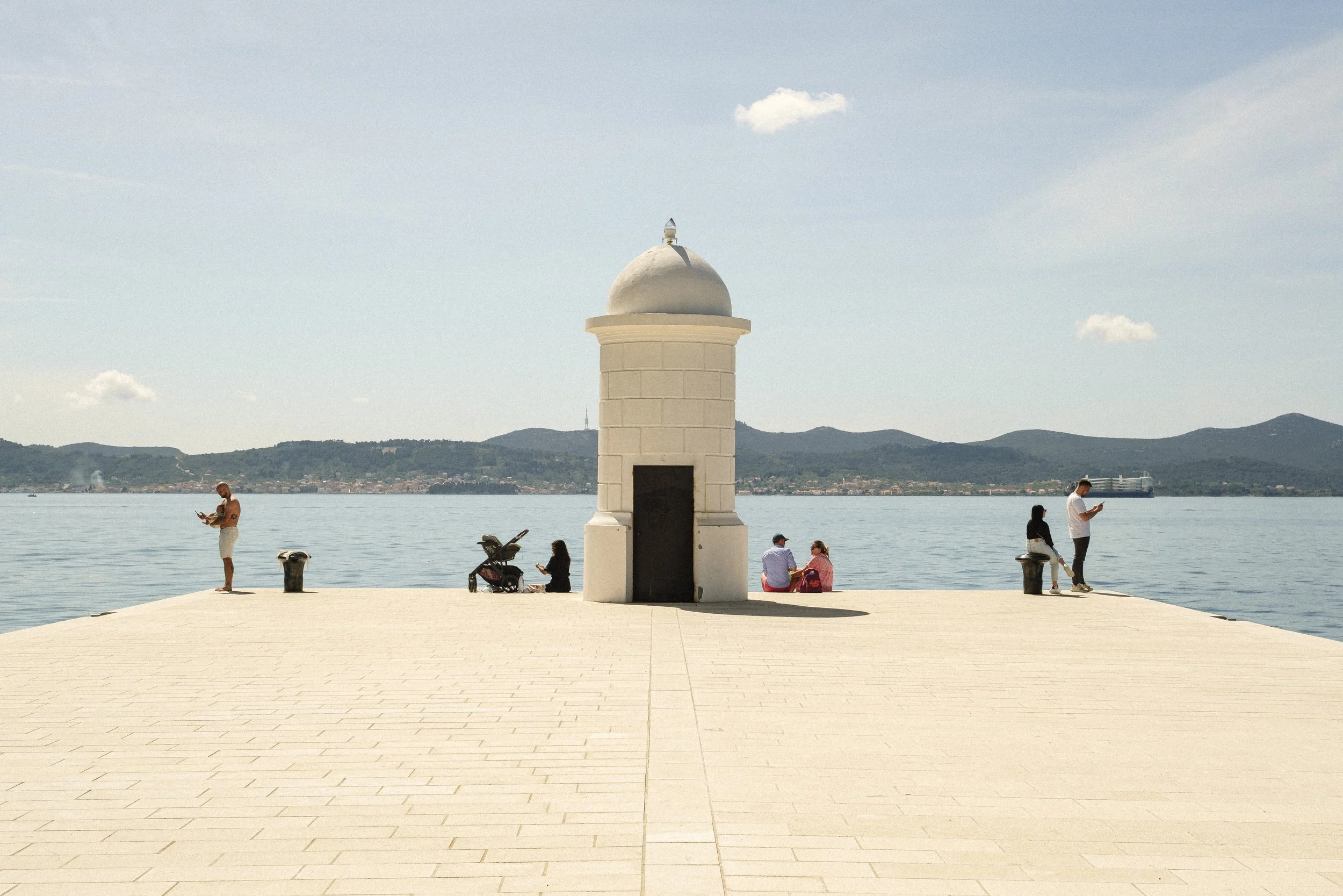 En liten fyr vid vattnet med fyra personer som sitter eller står i närheten, och några fler som är upptagna med sina telefoner.