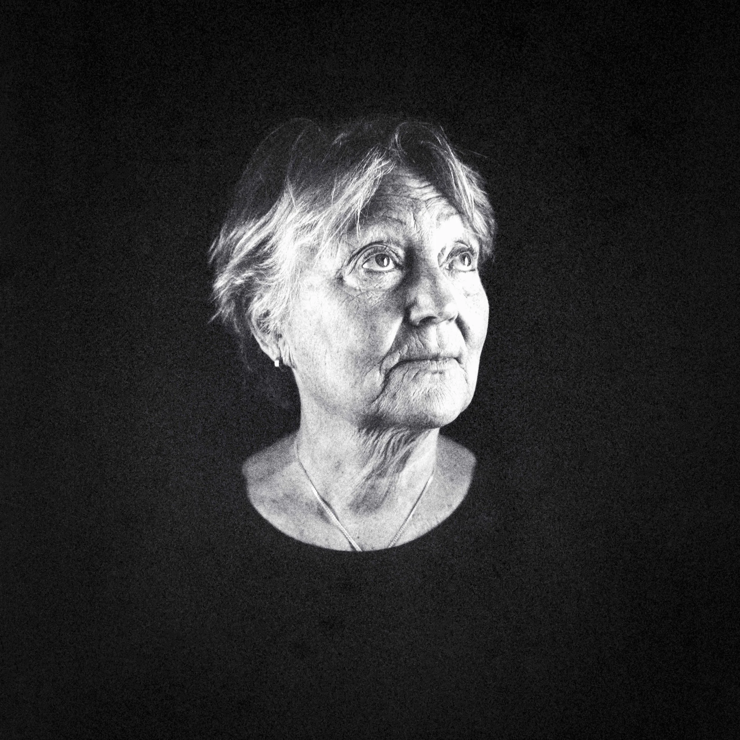 Svartvit porträttbild av en äldre kvinna med grått hår, tittande uppåt, mot en mörk bakgrund.