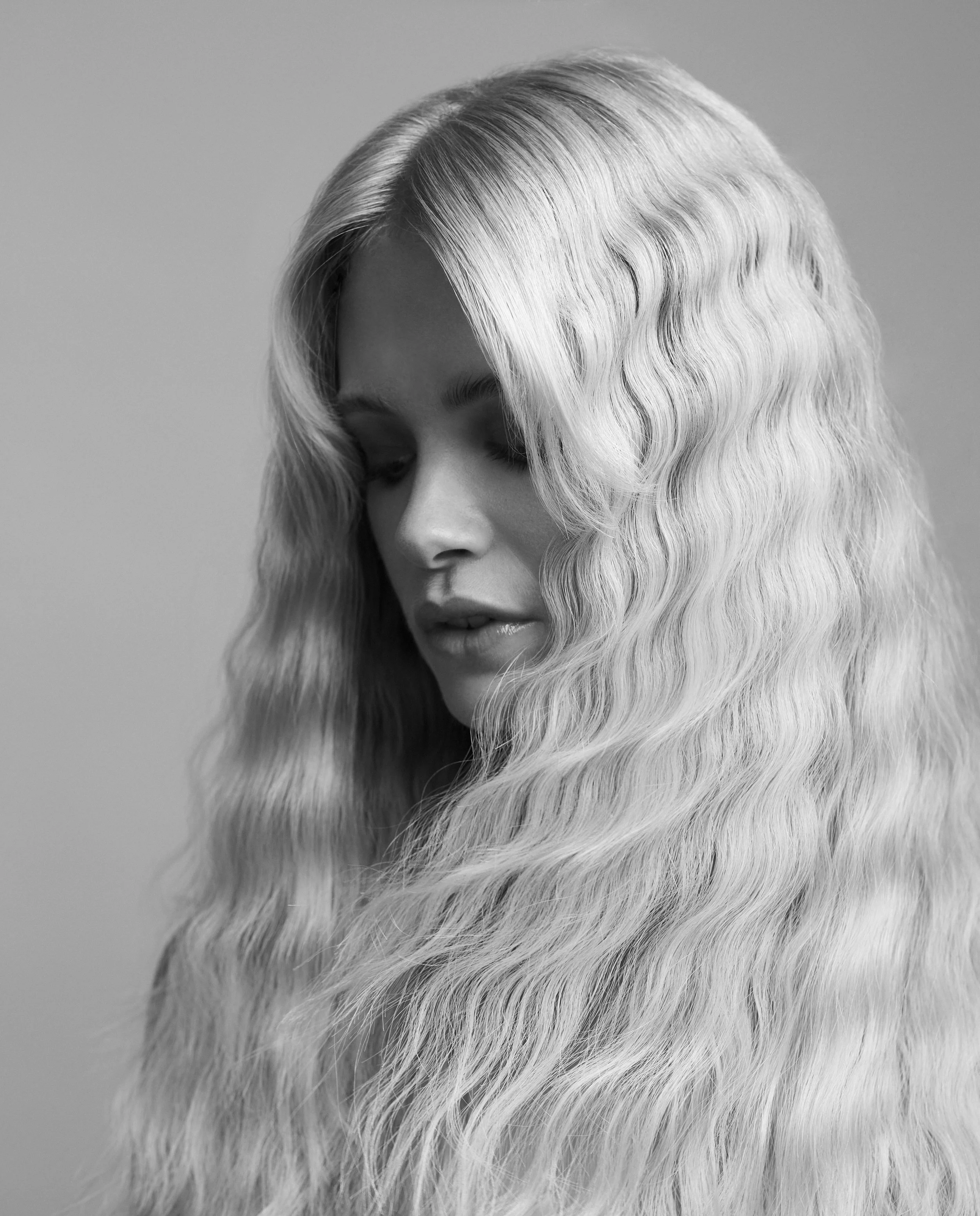 A woman with long, wavy, platinum blonde hair looking downward, in black and white.