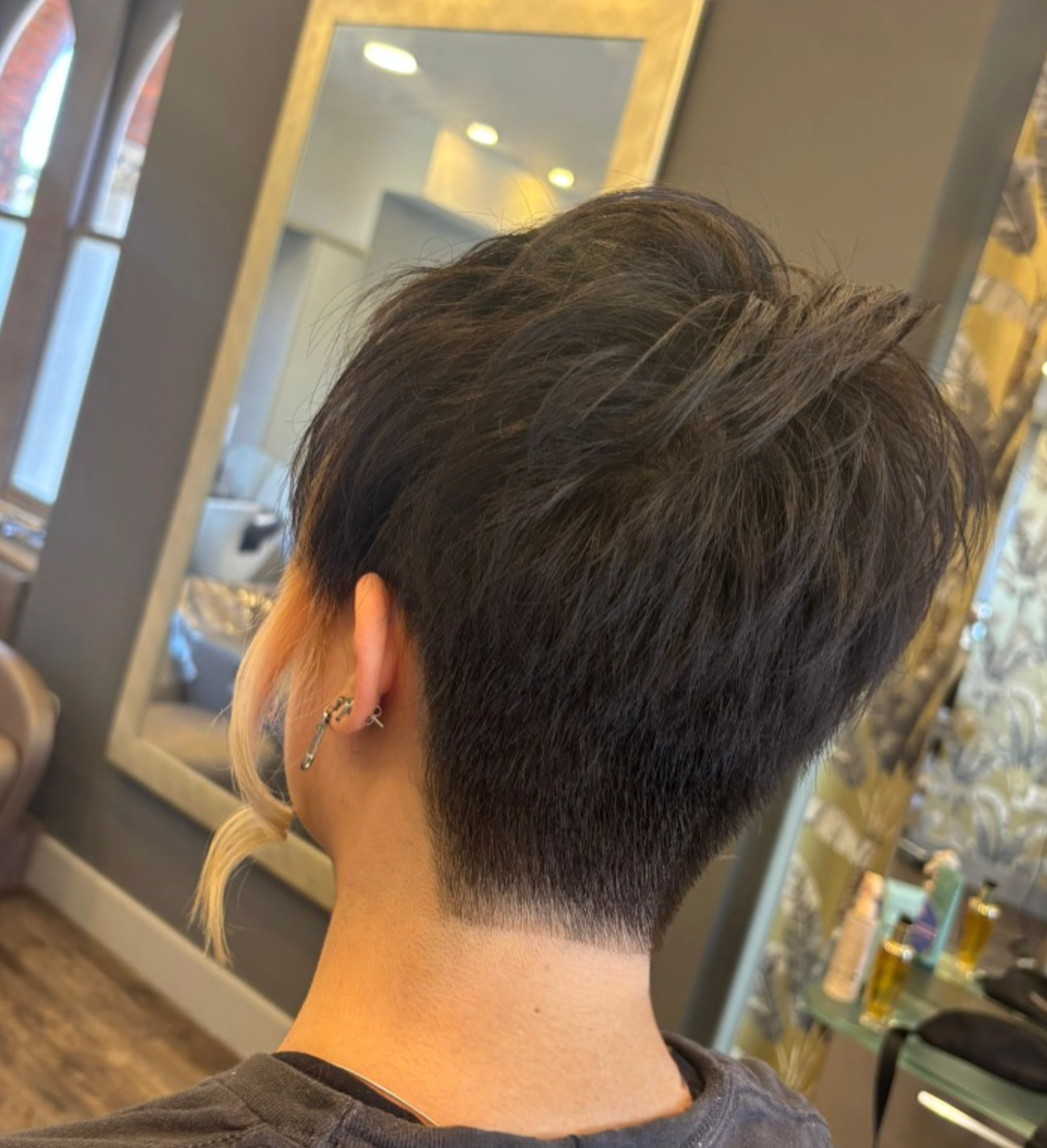 Back view of a person with a short, layered haircut with tapered sides and longer textured hair on top, in a salon setting.