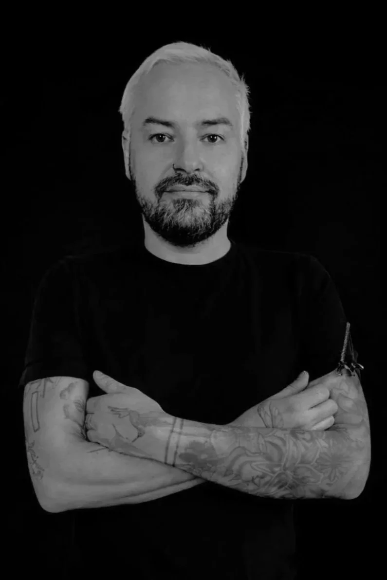 A smiling man with a beard and nose piercing, wearing a dark sweatshirt, sitting in front of a wall with banana leaf patterns.
