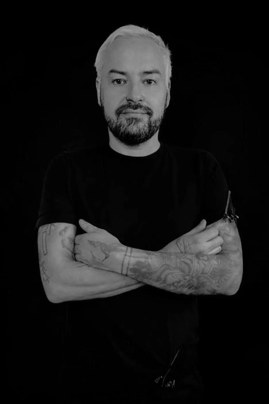 A man with a beard and mustache smiling, sitting in front of a wall with a tropical leaf pattern.