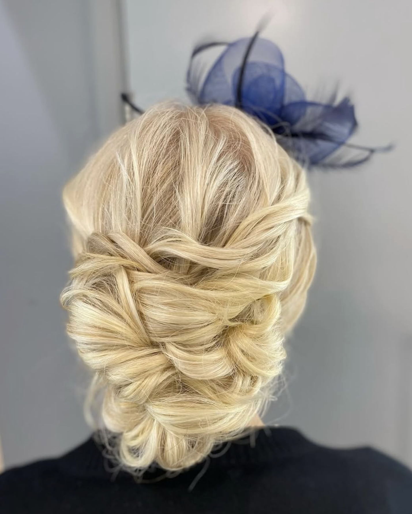 Blonde hair styled in an elegant updo with twists and loops, seen from the back, with a decorative blue hat with feathers in the background.
