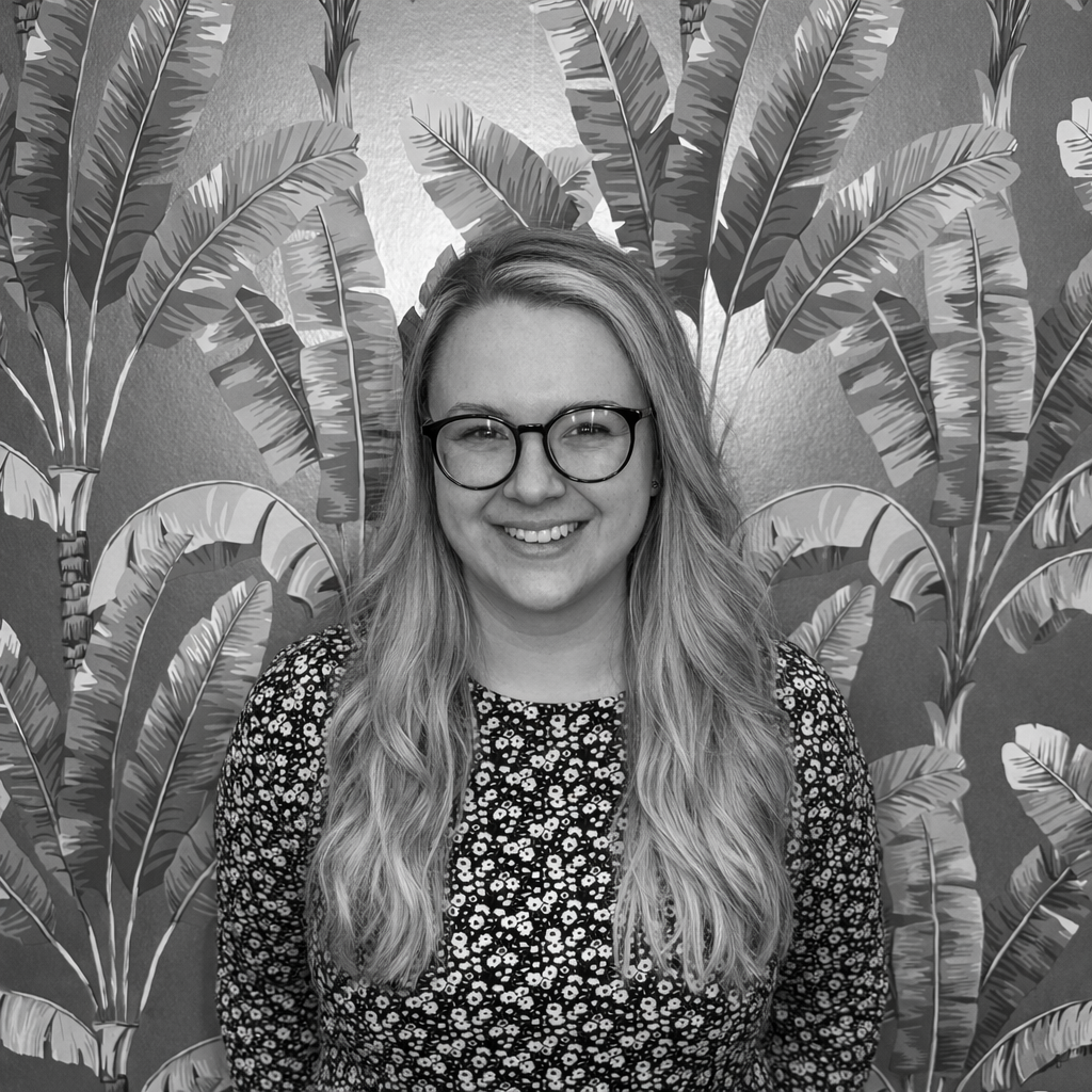 A woman with long wavy hair, glasses, and a floral patterned top, smiling and standing in front of a wall decorated with banana leaf patterns.