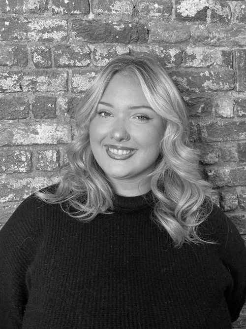 Black and white photo of a woman with wavy blonde hair, smiling, standing in front of a brick wall.