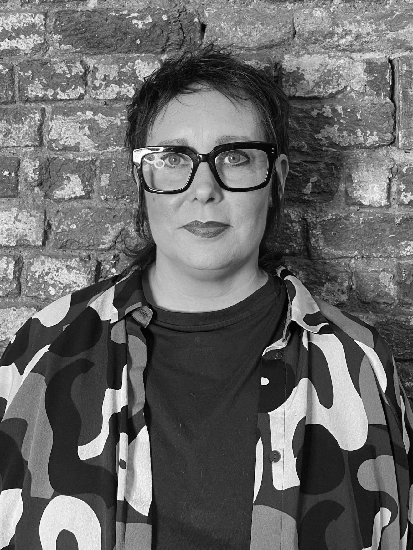 Black and white photo of a woman with short hair, wearing large eyeglasses, looking at the camera with a serious expression in a room with a chandelier and furnishings.