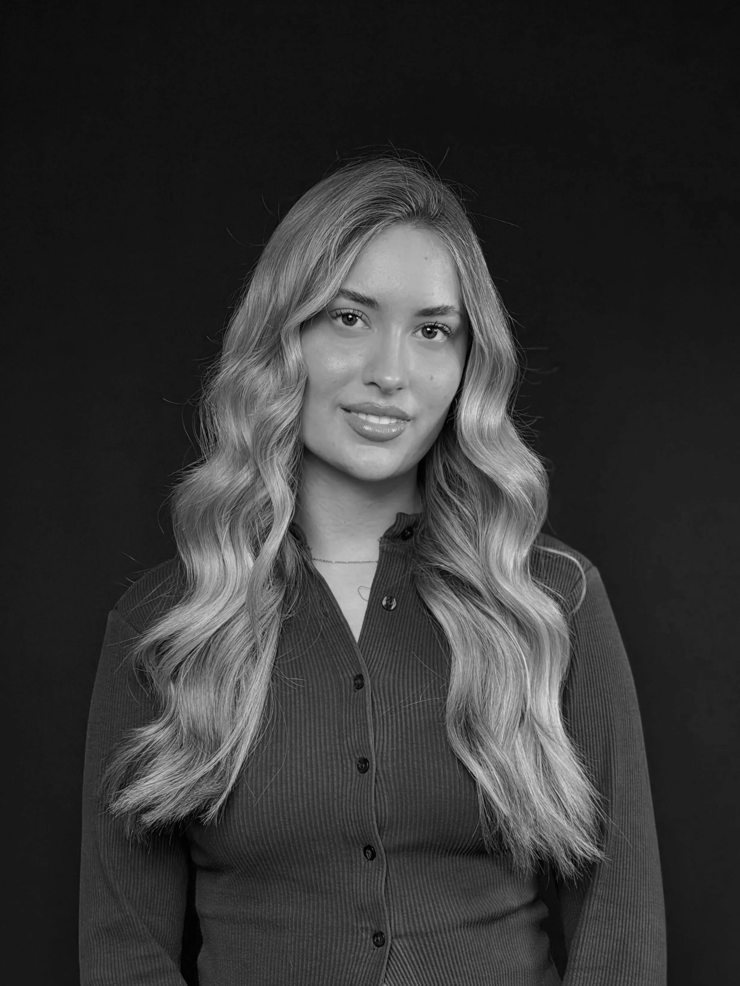 A woman with long hair and a slight smile standing in front of a tropical leaf patterned wall.