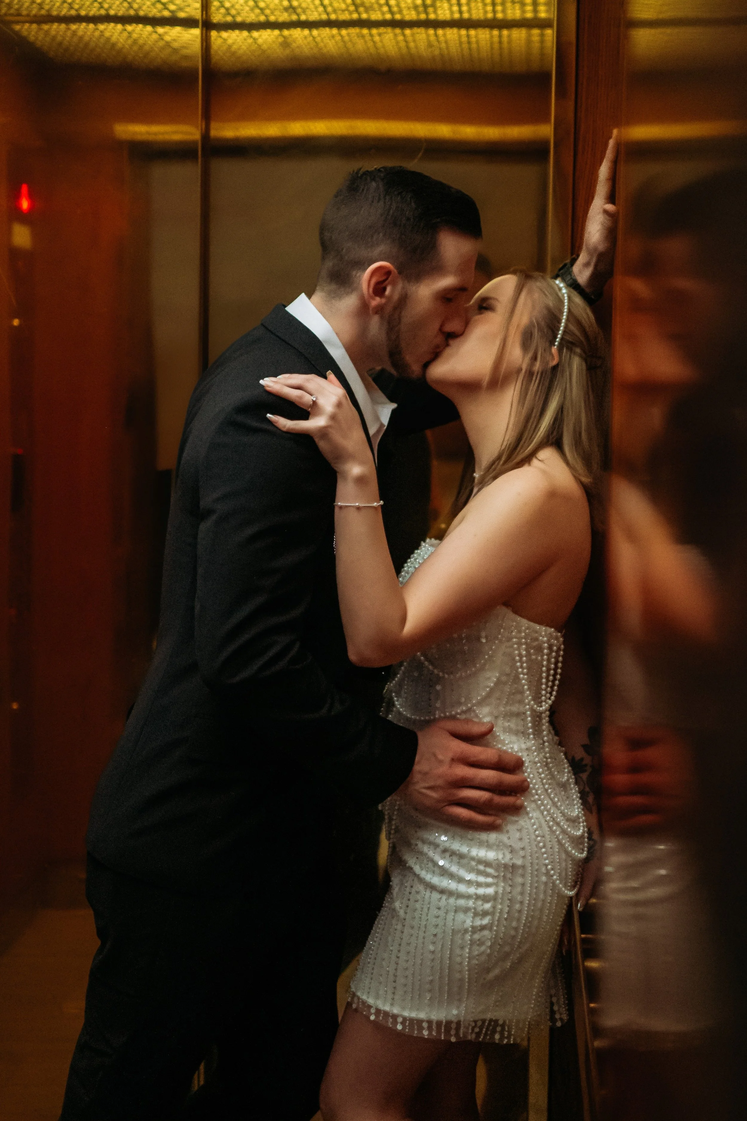 A couple kissing inside an elevator, with the man wearing a black suit and the woman in a pearl-studded white dress, leaning against a wall.