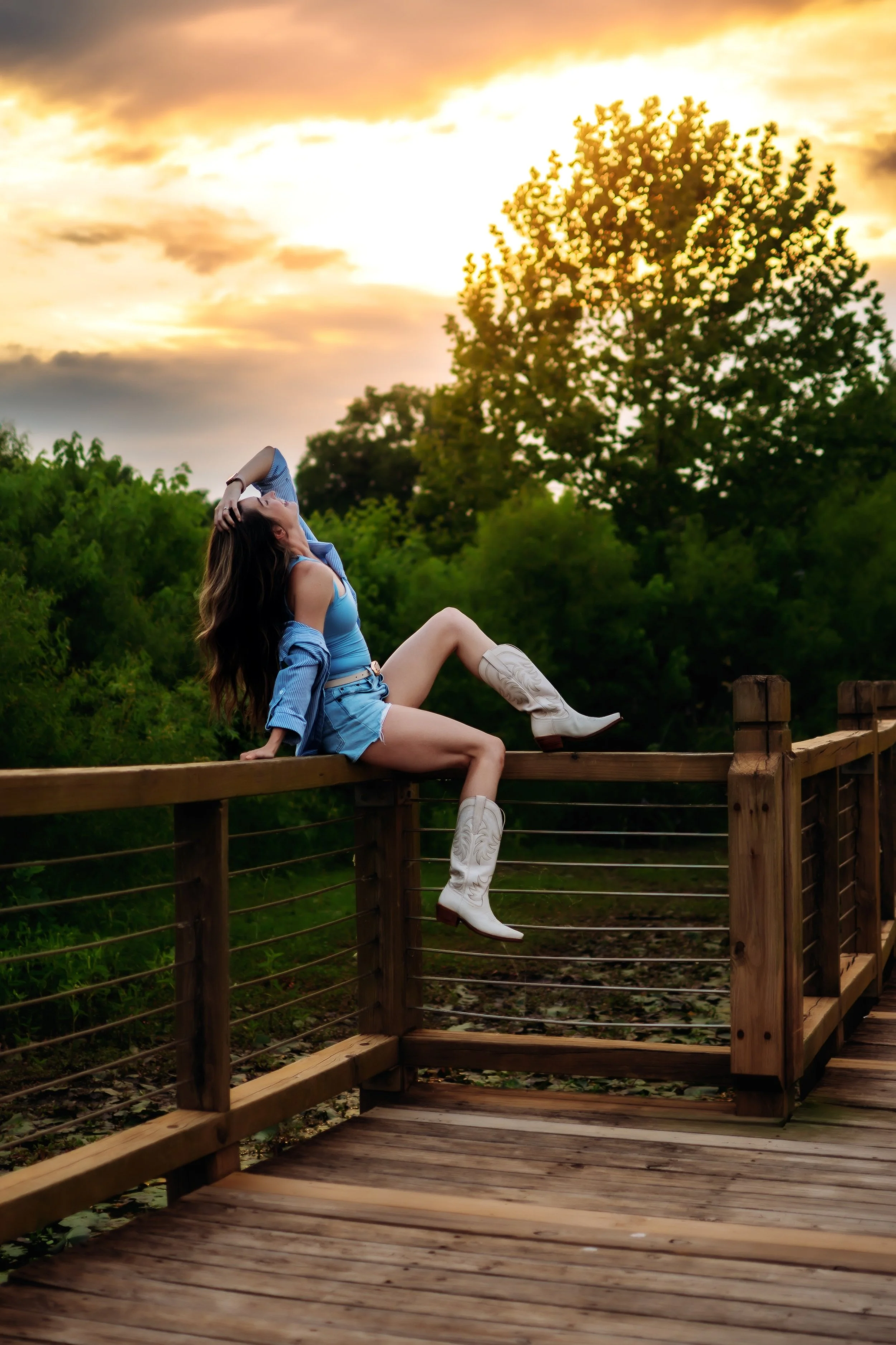 A woman wearing a blue crop top, shorts, and white cowboy boots sits on a wooden railing outdoors during sunset, with trees and a cloudy sky in the background.