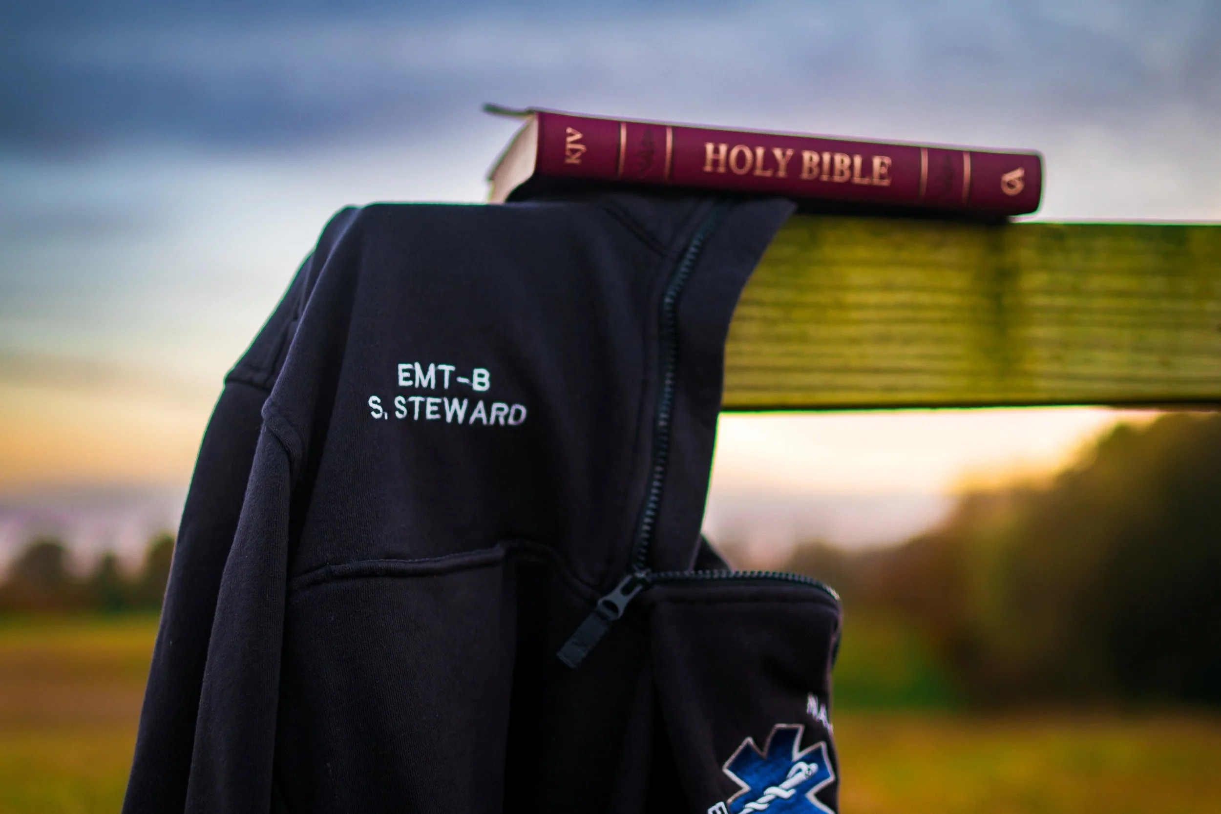 EMT work jacket laying on the edge of a cross with the Holy Bible on top. Sunset in the background
