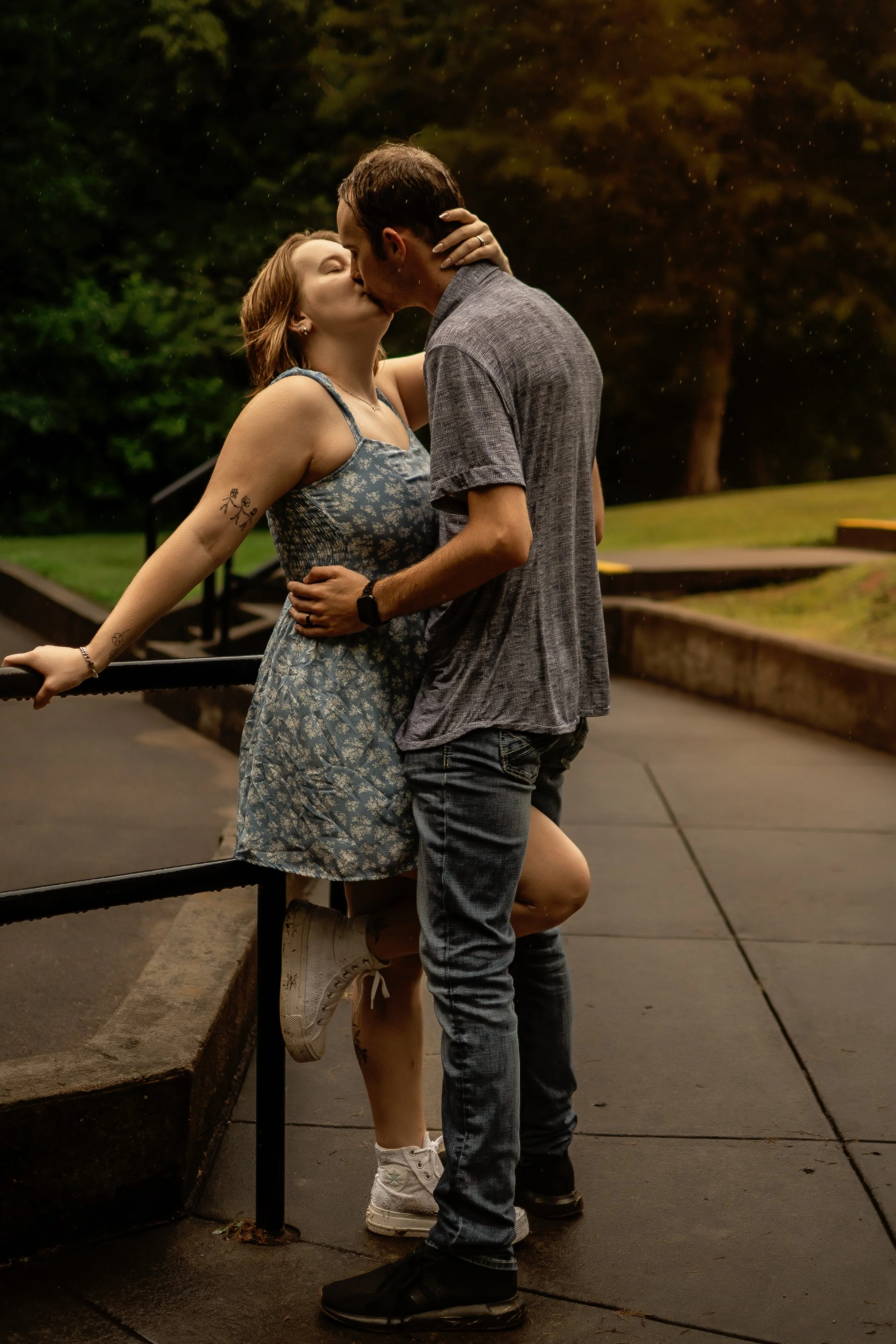 A couple sharing a passionate kiss outdoors at night, with a woman leaning on a railing and a man standing close to her, both dressed casually.