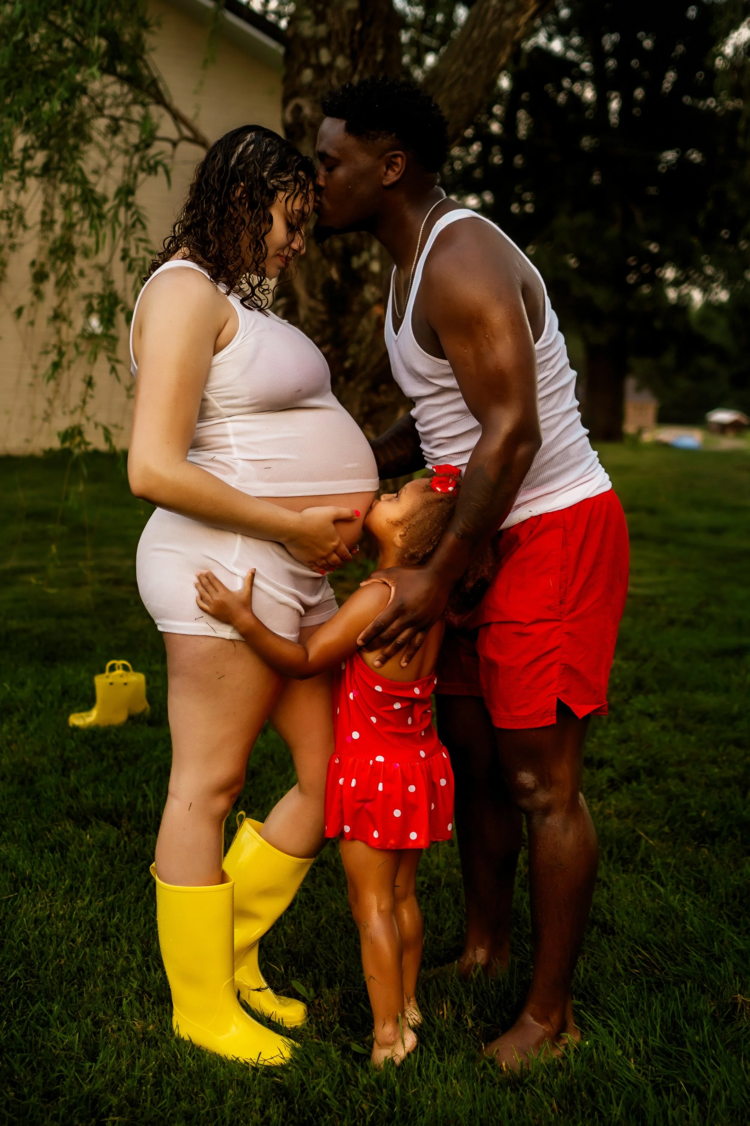 A pregnant woman and a man are standing close together outside, touching foreheads and holding a young girl, all in casual summer clothing. The woman wears a white tank top and shorts with yellow rain boots, the man is in a white tank top and red shorts, and the girl is in a red polka-dot dress with yellow boots. They are in a grassy yard with a tree behind them.