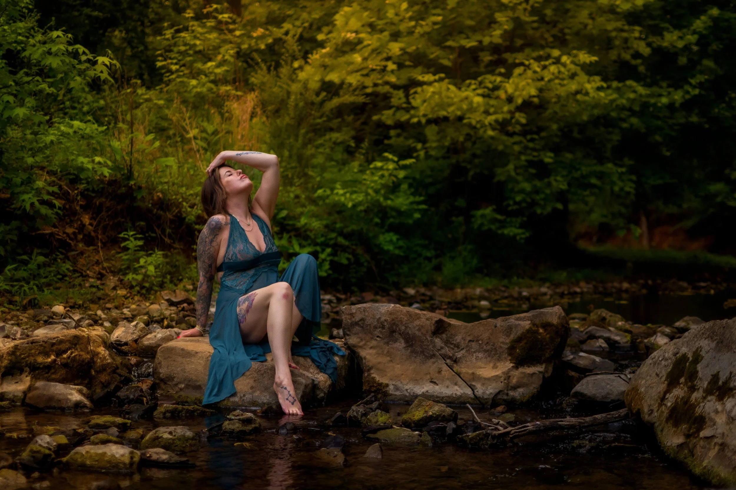 women in a sheer blue outfit sitting on a rock in a creek with one hand above her in her hair