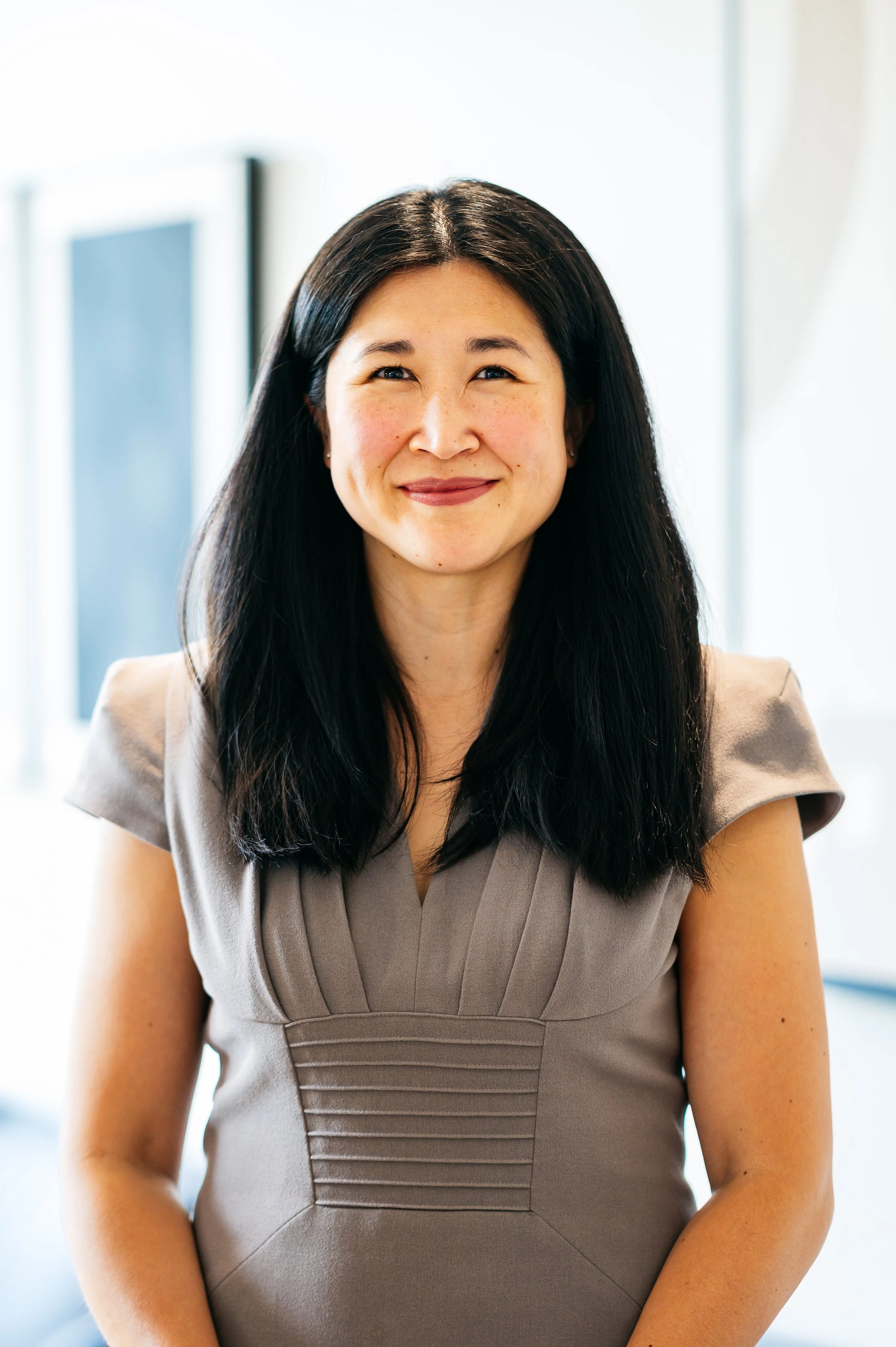 A smiling Asian woman with long black hair wearing a black sleeveless top and gold earrings against a black background.