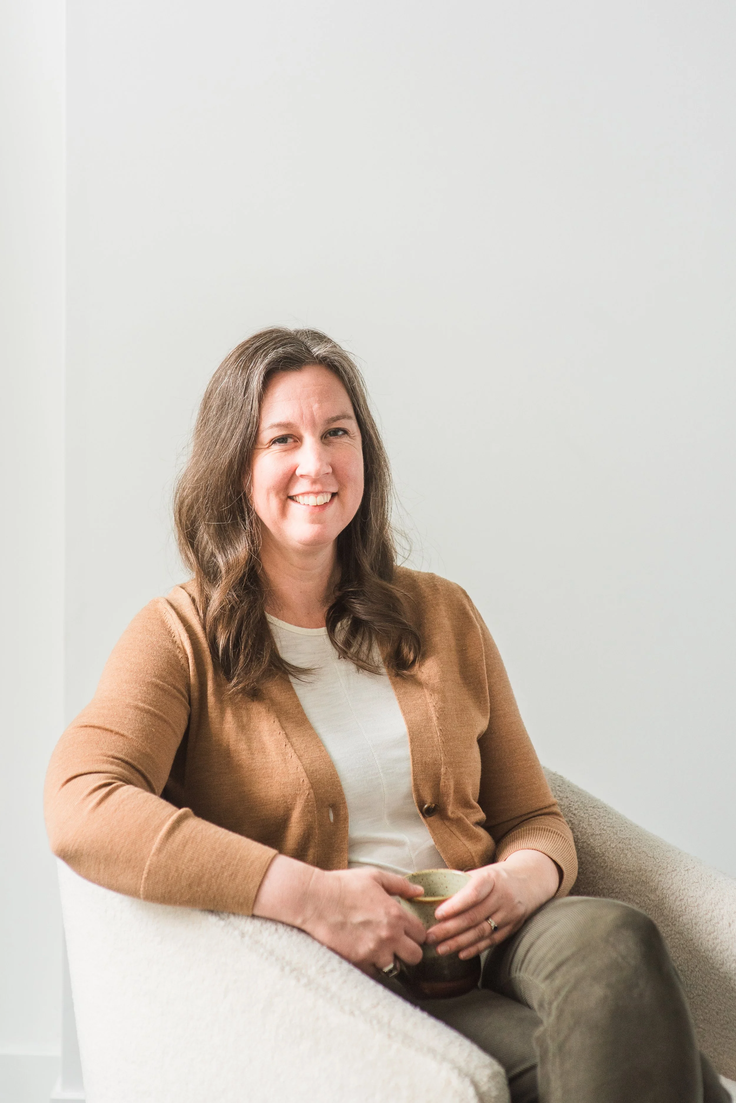 A woman with brown hair smiling and sitting on a beige armchair, holding a cup, wearing a tan cardigan and a white top, against a plain light-colored wall.