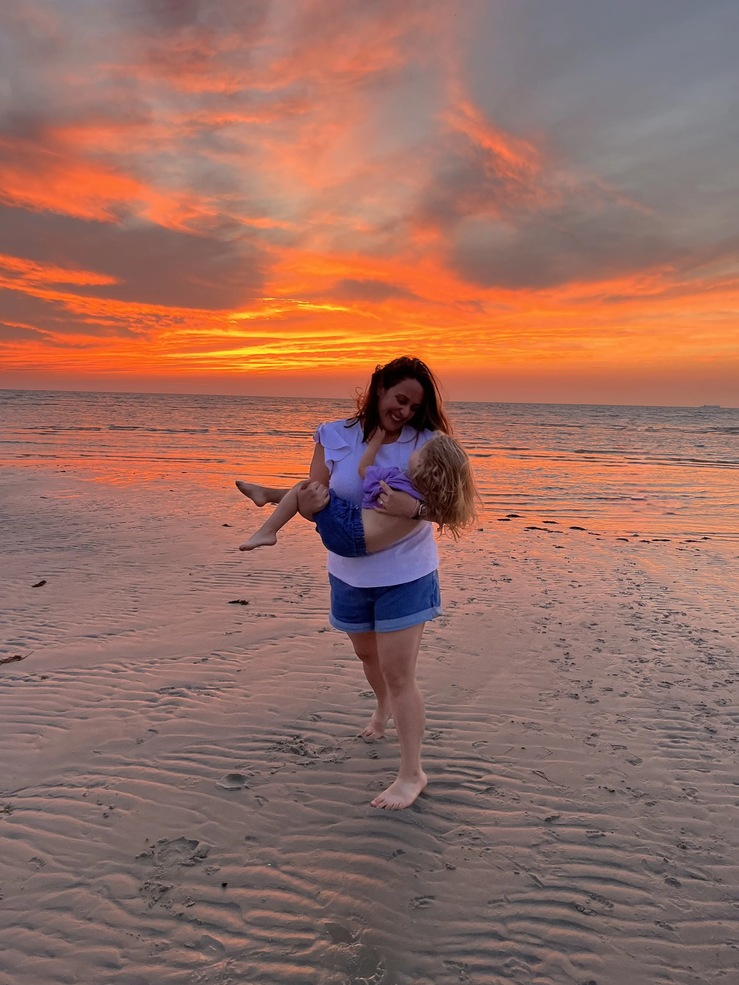 A mother with curly hair is holding her young daughter  with blonde hair on a beach during a colorful sunset, both smiling and enjoying the moment.