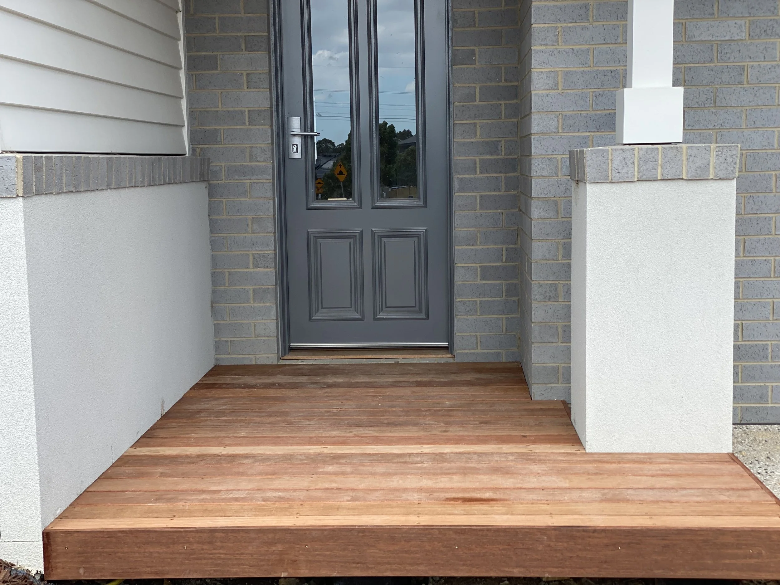 Front porch with wooden deck in front of a gray door, brick walls, and white column on the right.