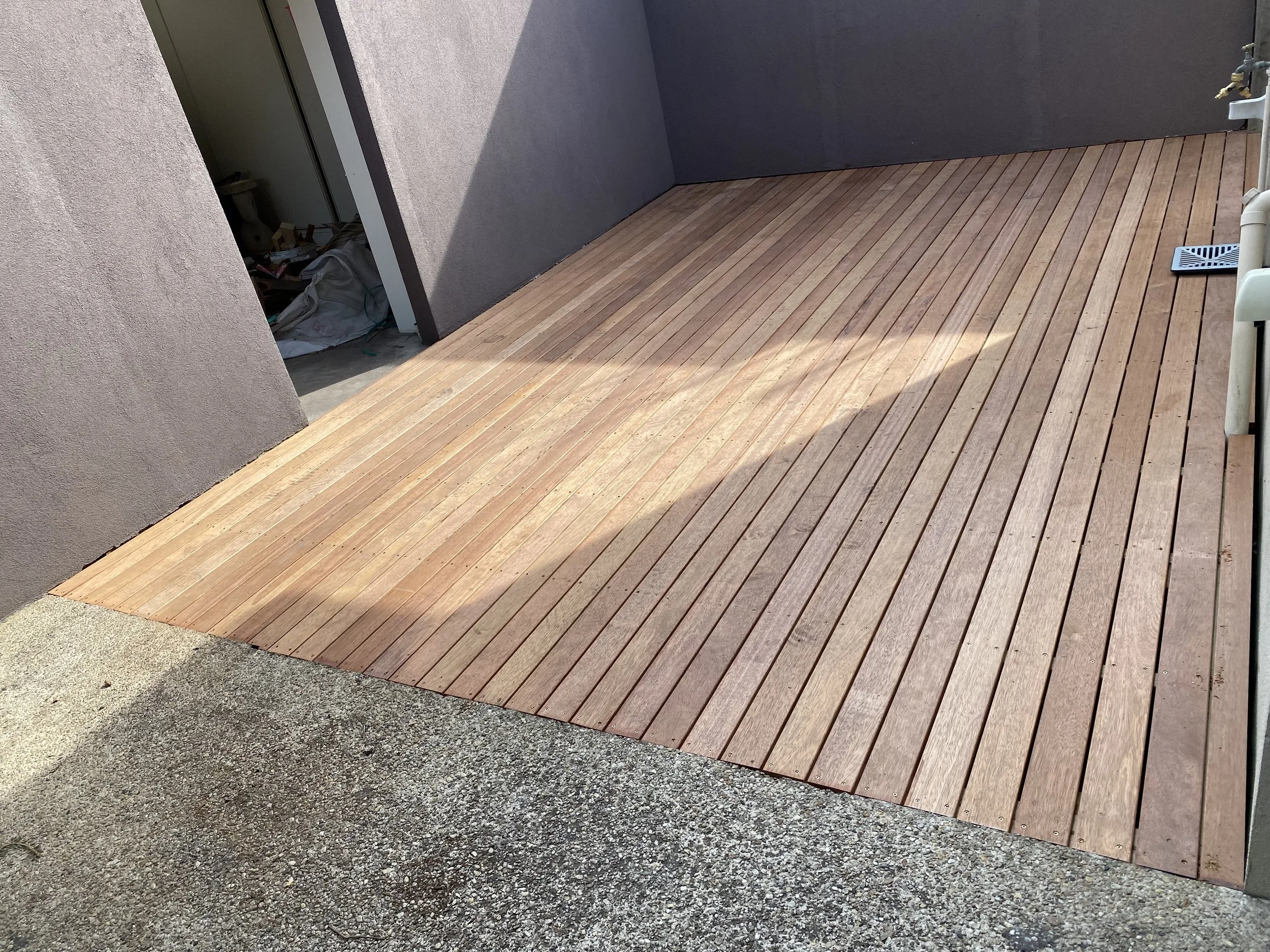 Newly installed wooden deck outside a building, with an area of rough concrete flooring and an open door leading inside.