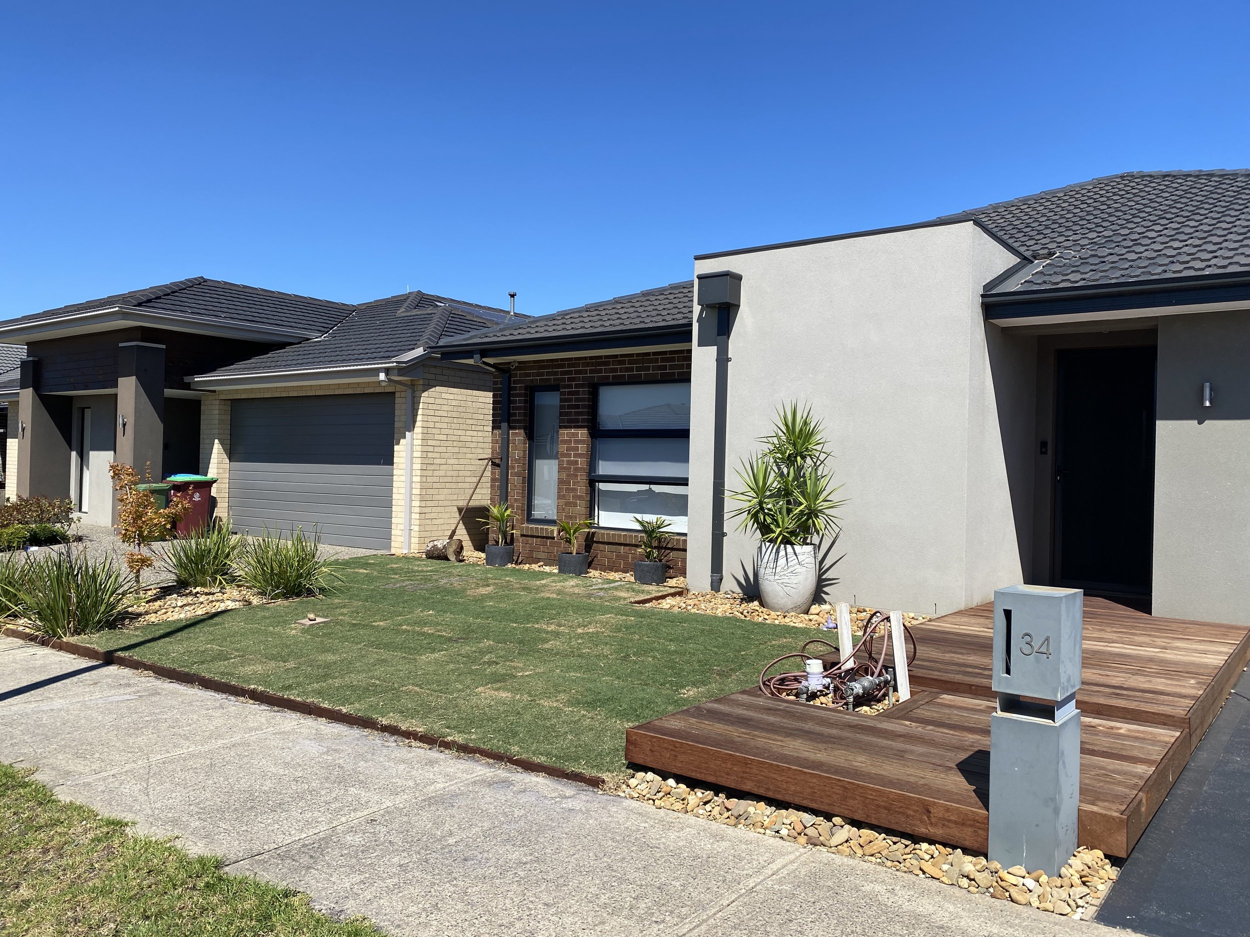 Front view of modern suburban house with white and brick exterior, small front yard with grass and plants, wooden porch area with a mailbox numbered 134, and blue clear sky in the background.