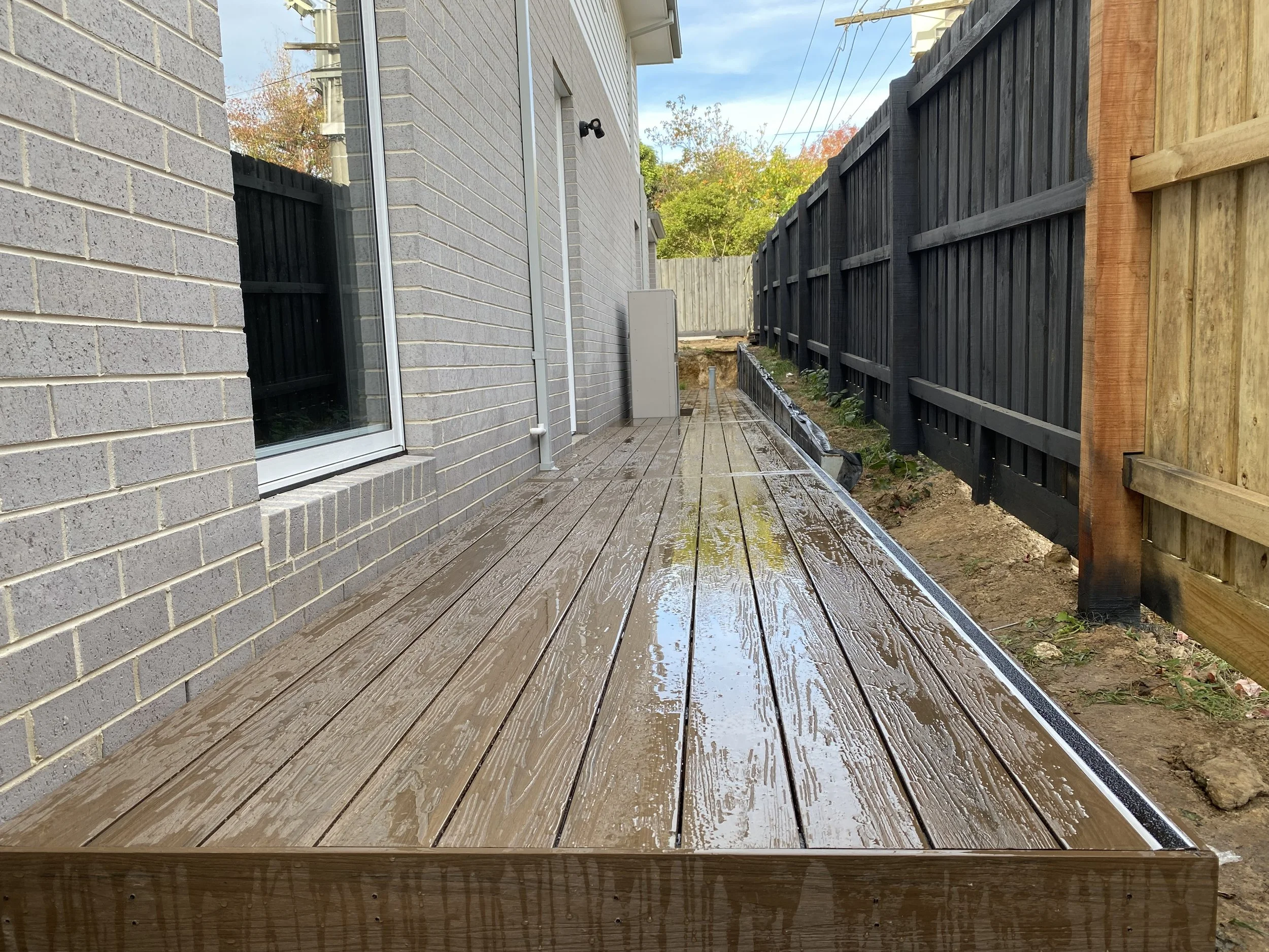 Wet wooden deck outside of a house with gray brick siding, a window, and a black wooden fence on the right side. Some trees are visible in the background.