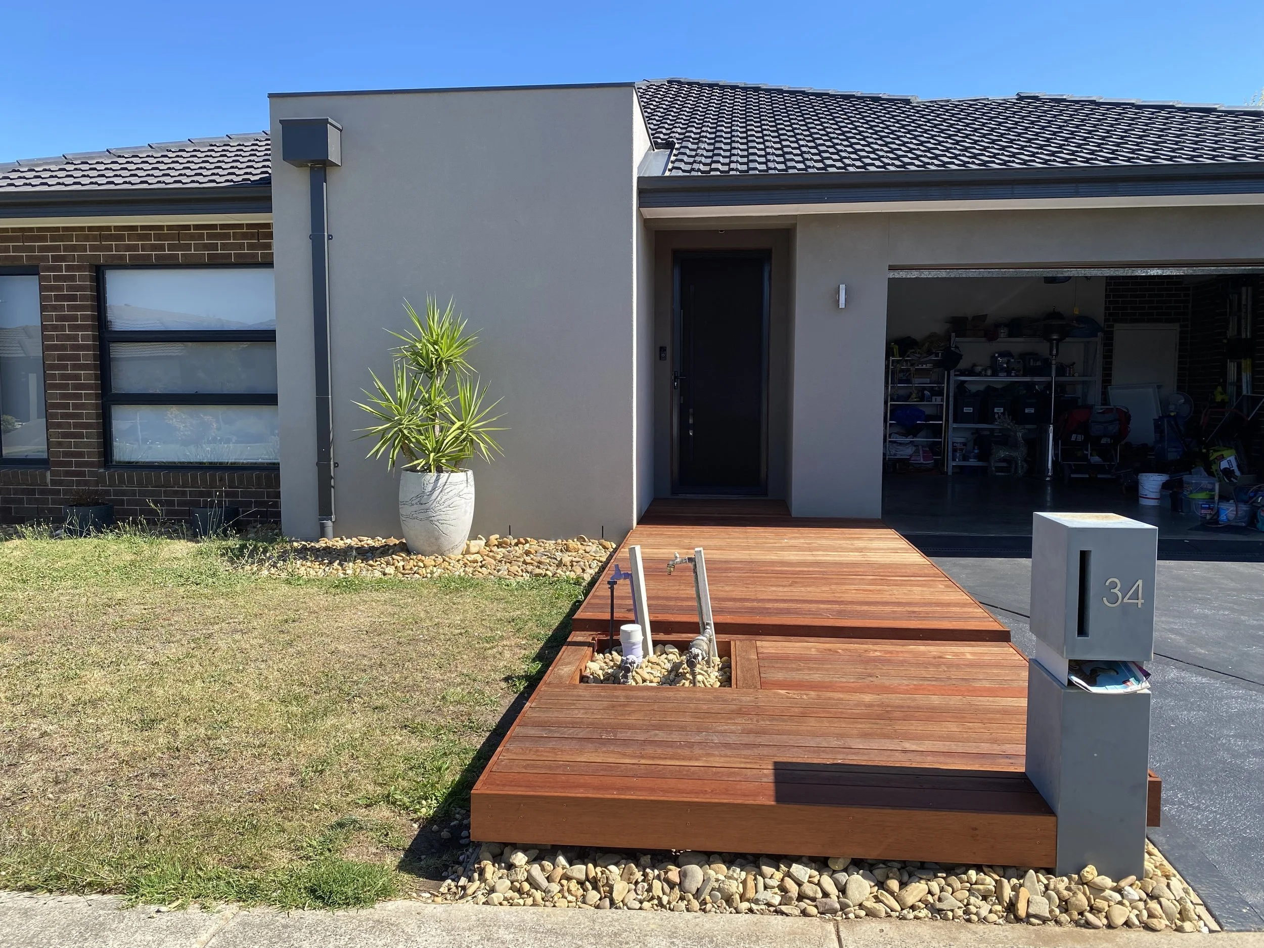 Modern house with a wooden front porch, gray exterior wall, potted plant, and open garage filled with stored items. House number 34 on a gray mailbox at the sidewalk.