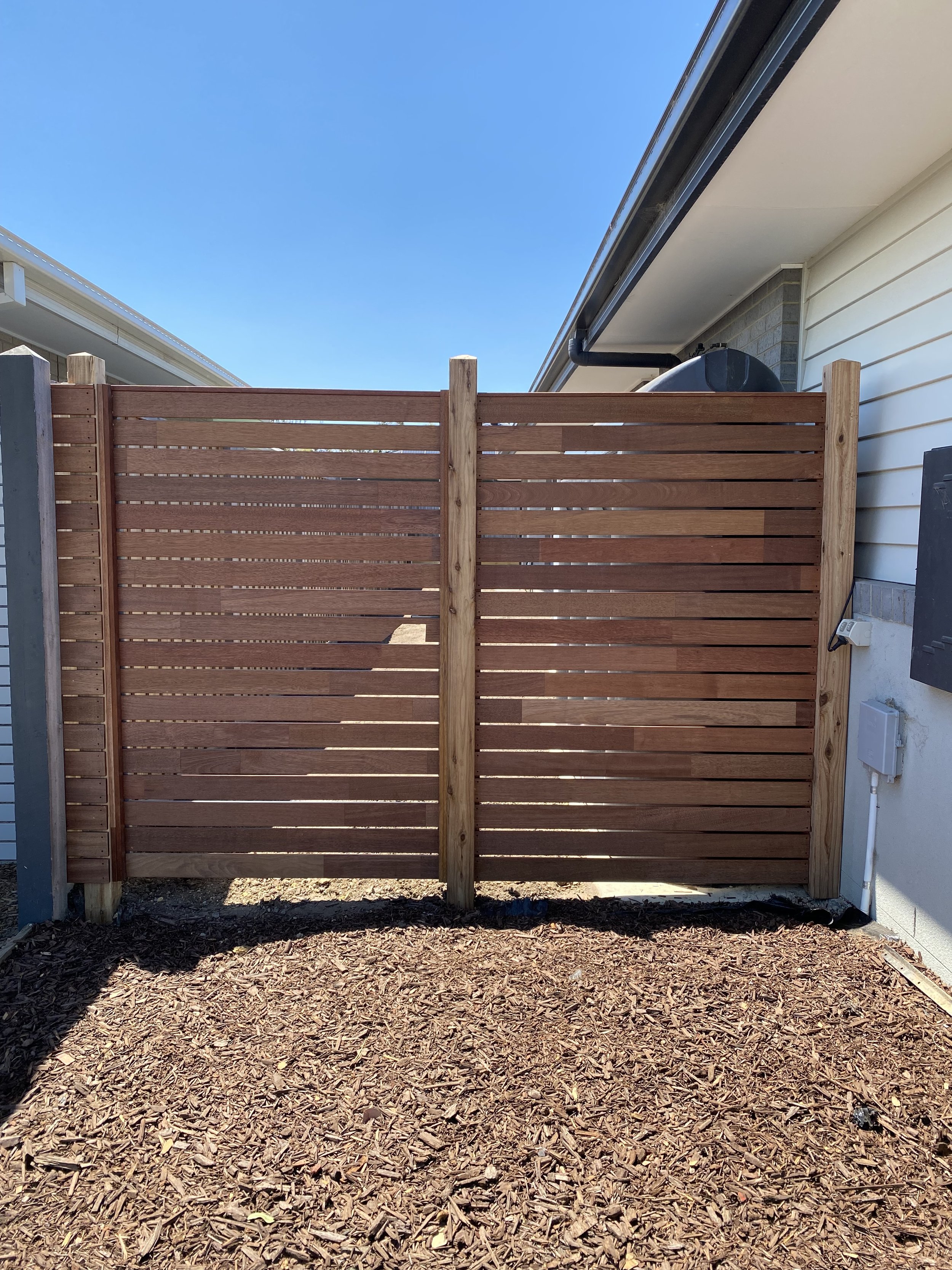 A wooden privacy fence with horizontal slats and vertical support posts, located between two houses with clear blue sky overhead.