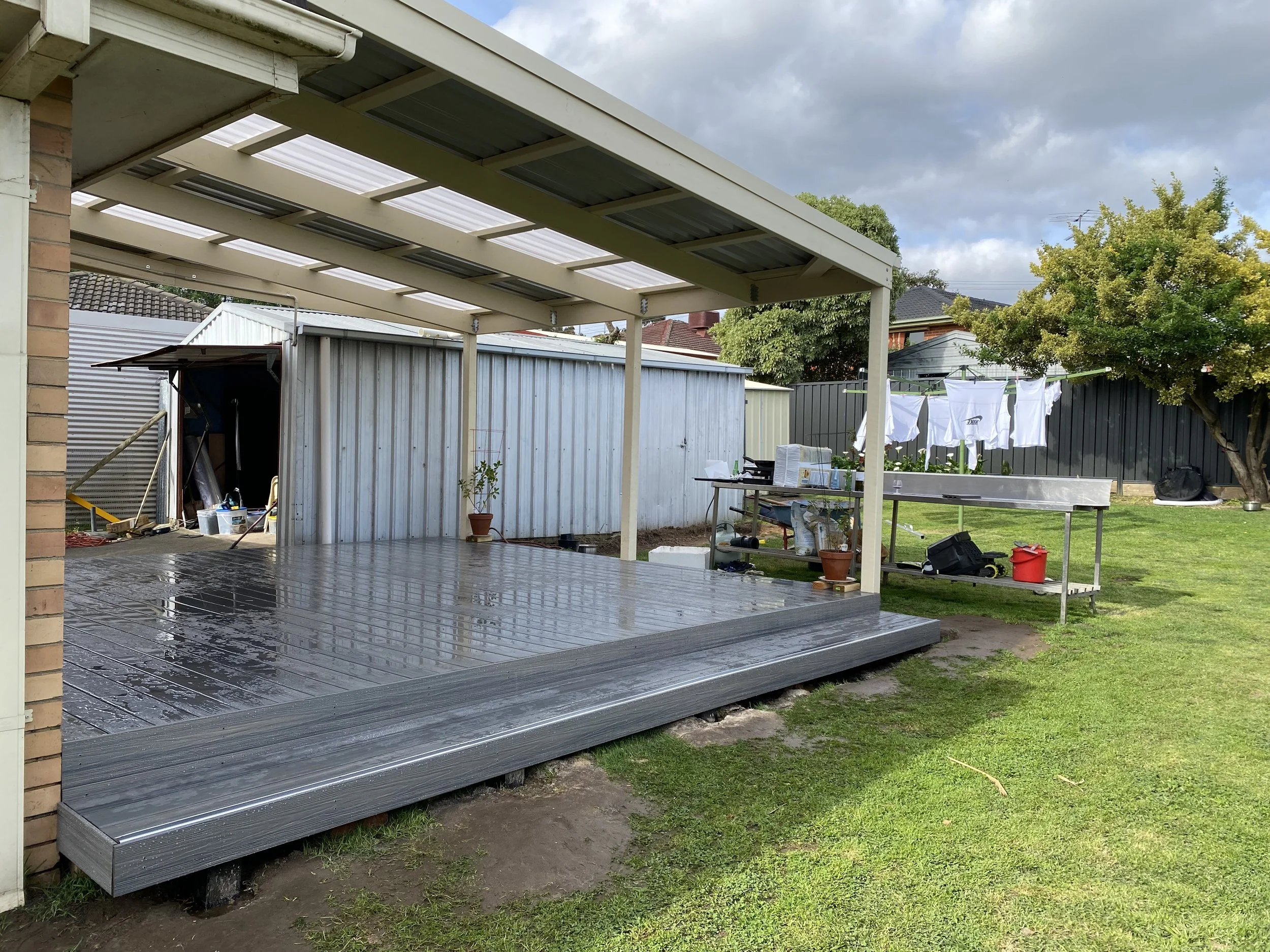 Backyard with partially covered outdoor deck, drying laundry on a clothesline, and gardening tools.