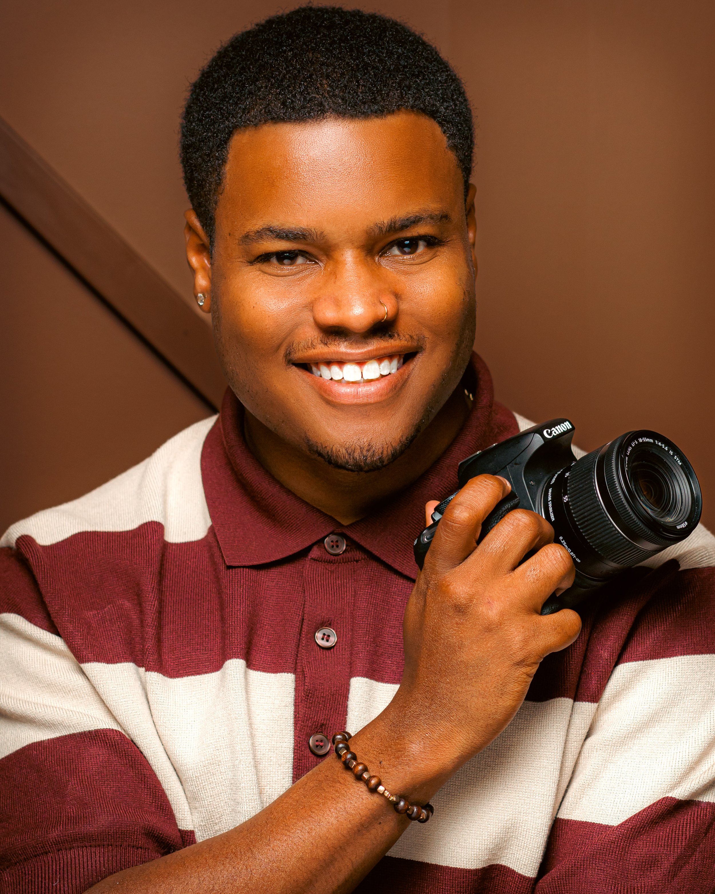 Close-up of a smiling person with short curly hair, wearing a striped sweater and holding a camera.