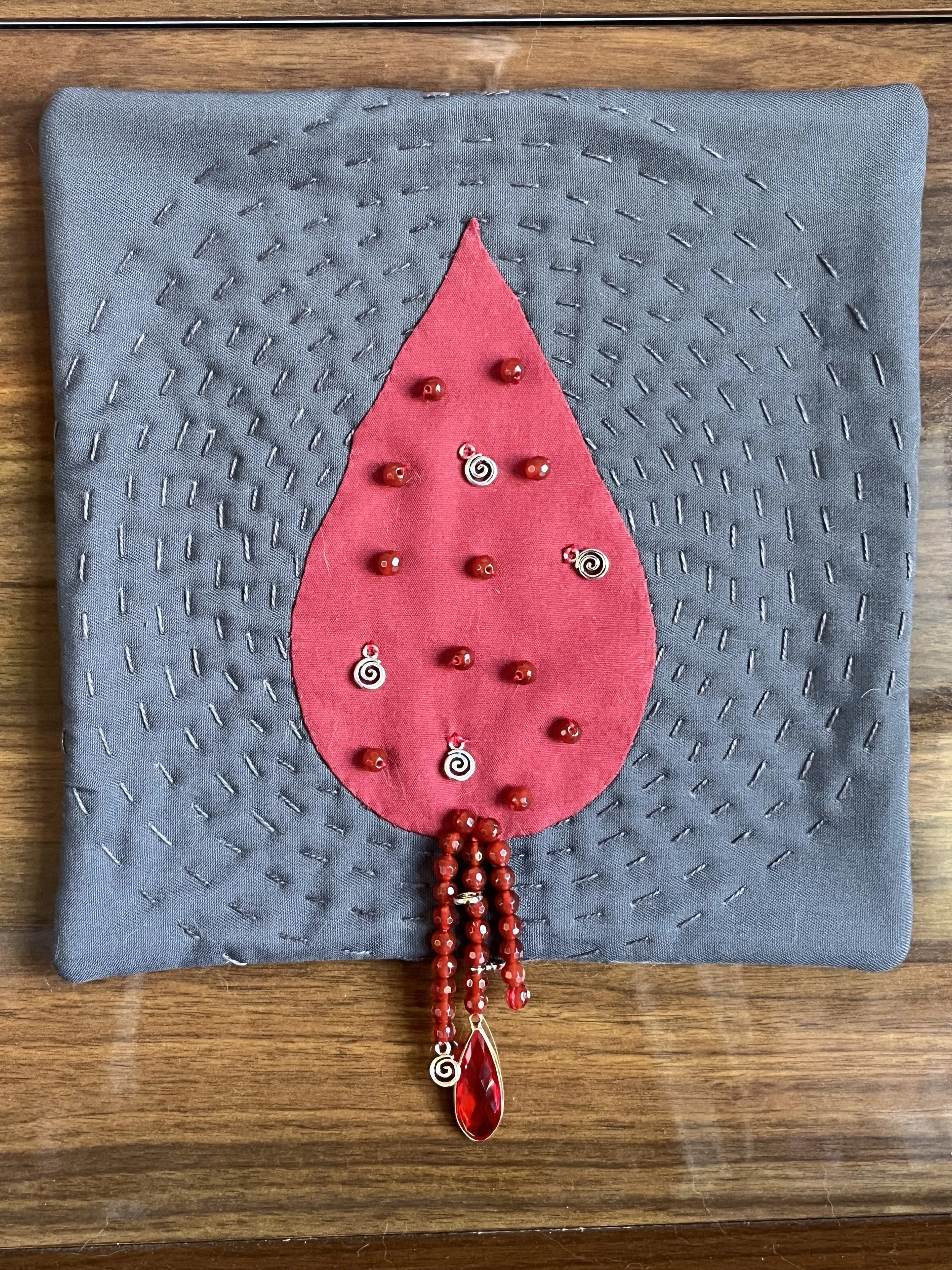 Small art quilt depicting a large drop of blood against a grey background. The drop is adorned with red agate beads and silver spirals.