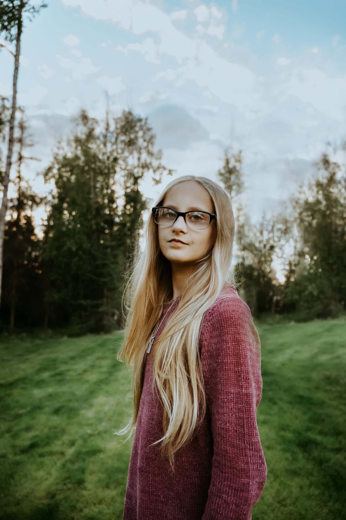 Young person with long hair and glasses standing on a grassy field with trees in the background.