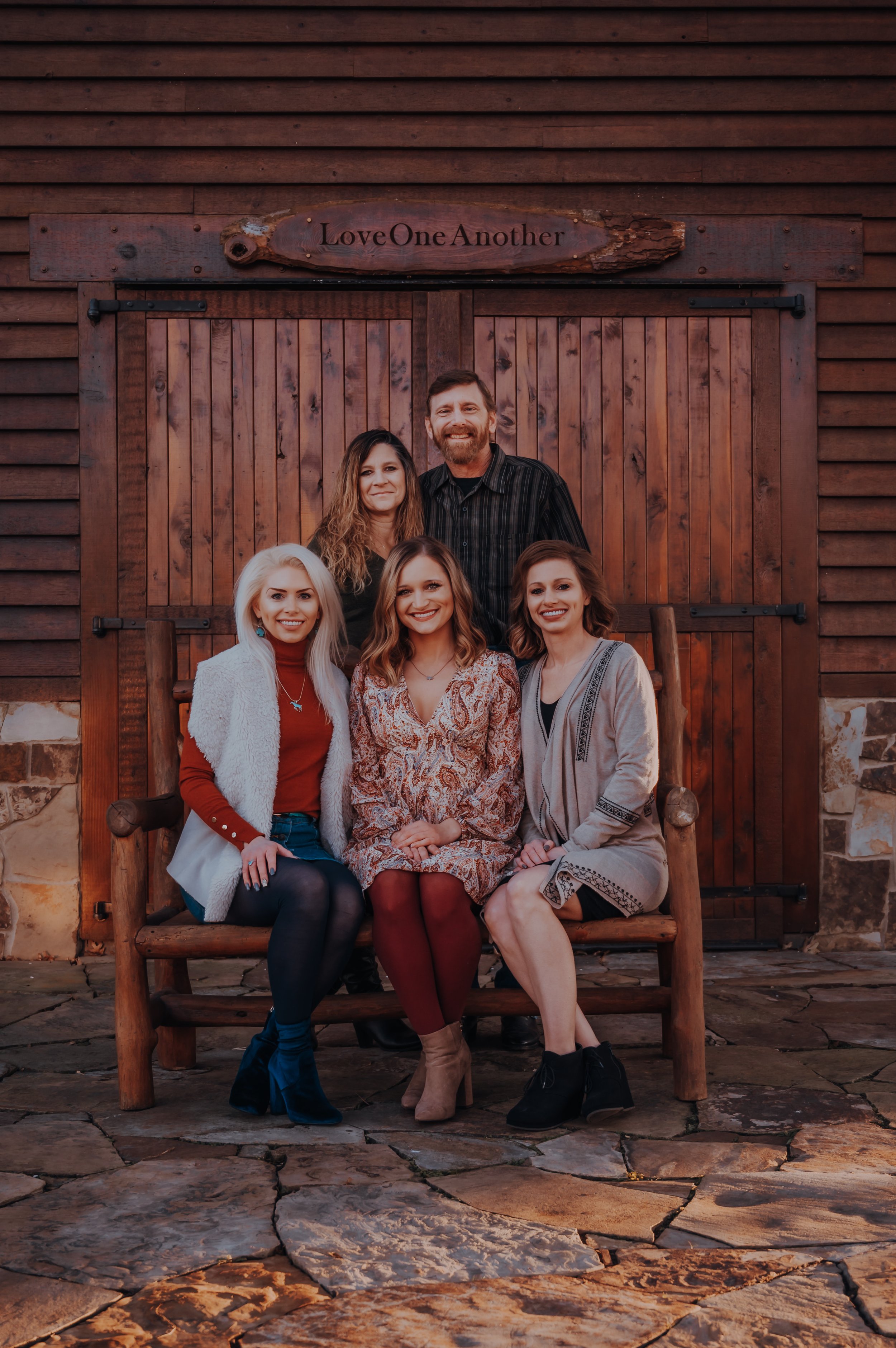 Group photo of three women seated and two women and a man standing behind them, with a rustic wooden door backdrop and a sign above reading 'Love One Another.'