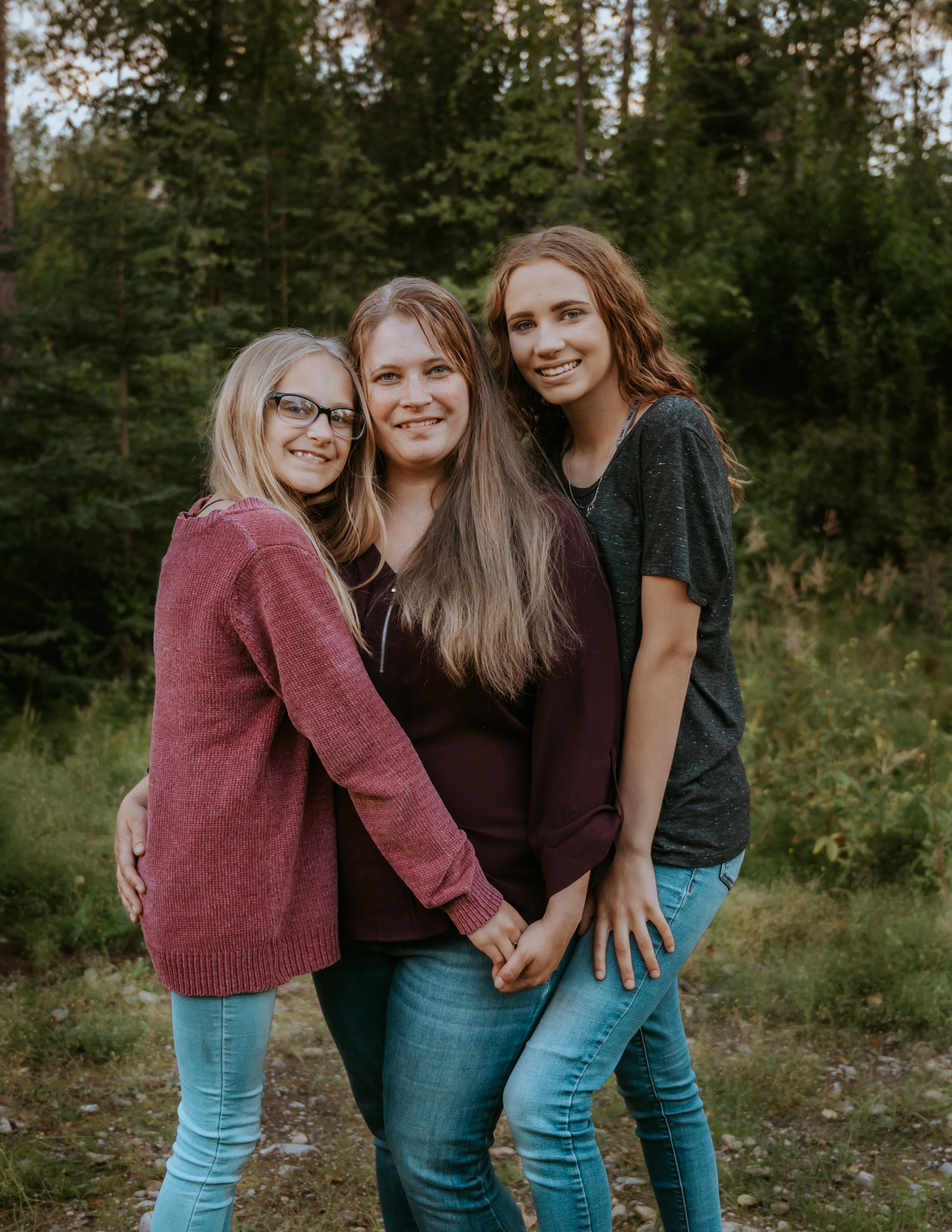 Three women standing close together, smiling in a forest setting, wearing casual sweaters and jeans.