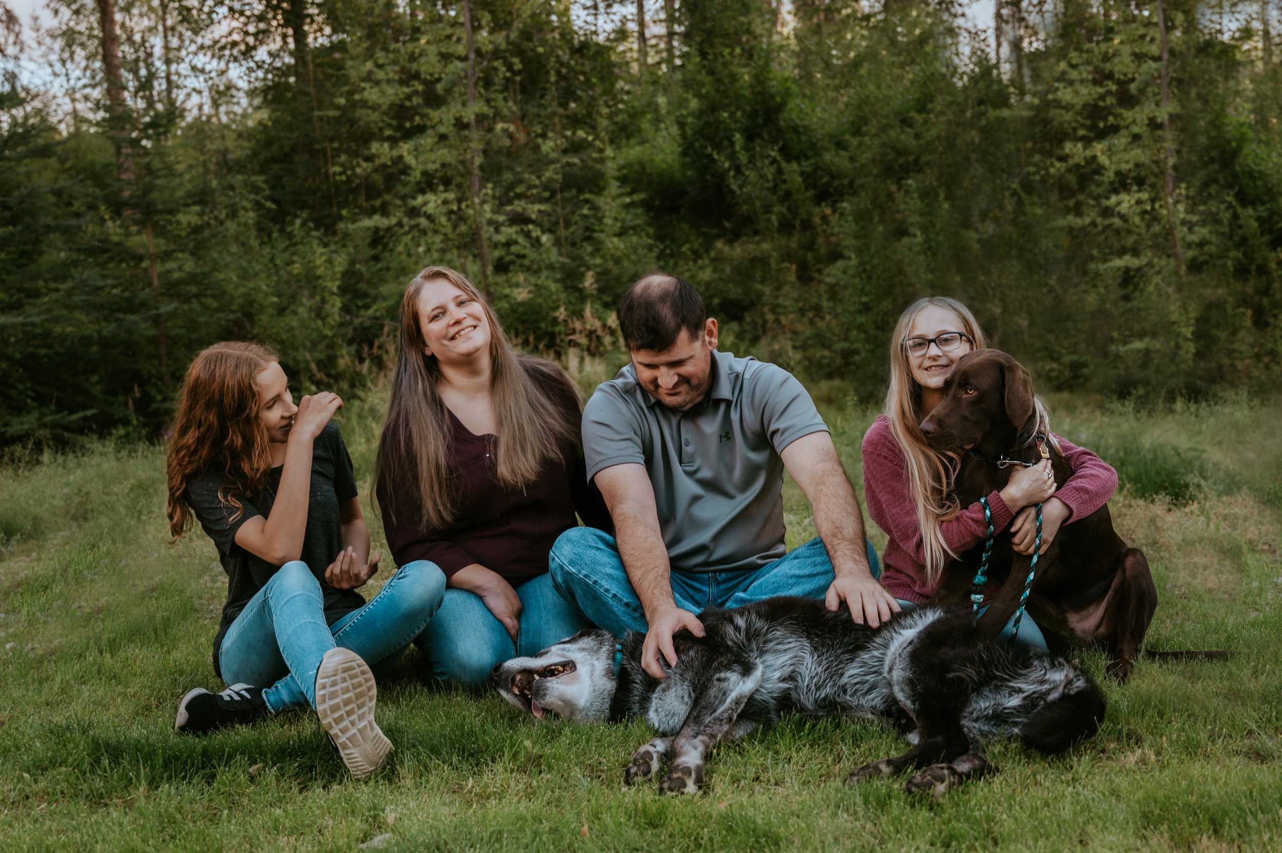 Family sitting on grass with two dogs in a wooded area.