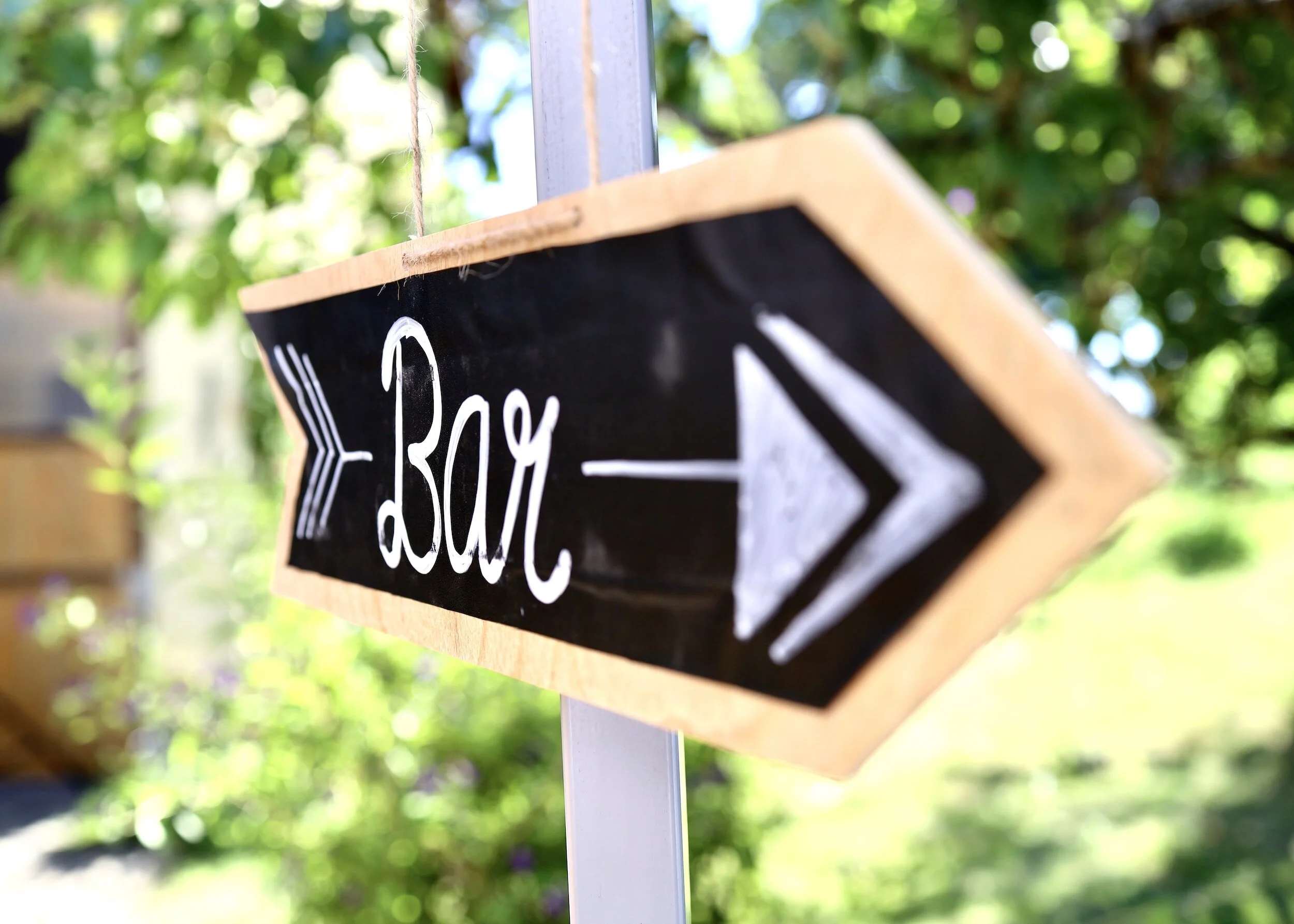 Close-up of a blackboard sign with white handwritten text and arrow, mounted on a wooden board, outdoor setting with blurred green foliage in the background.
